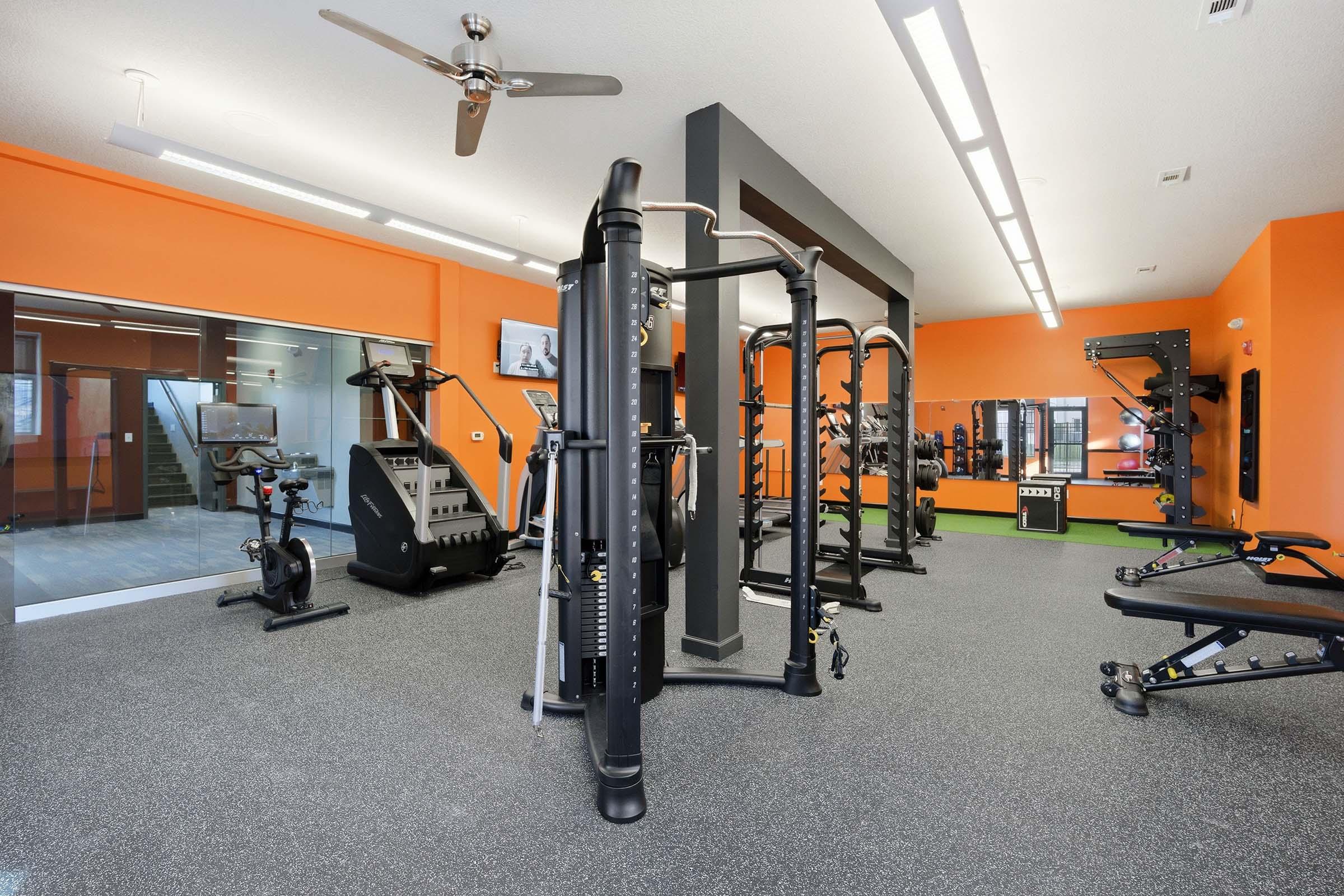 Modern gym interior featuring a variety of exercise equipment, including a weight machine, stationary bike, and benches. The walls are painted orange, providing a vibrant atmosphere. Large windows allow natural light to fill the space, creating an inviting workout environment.