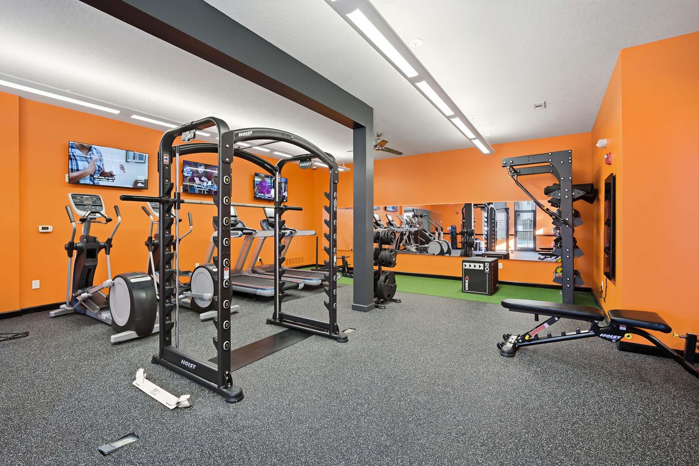 A modern gym with orange walls, featuring exercise equipment such as cardio machines, a squat rack, and a weight bench. Two wall-mounted TVs are visible, and a large mirror reflects the workout area. The floor is covered in black rubber matting.