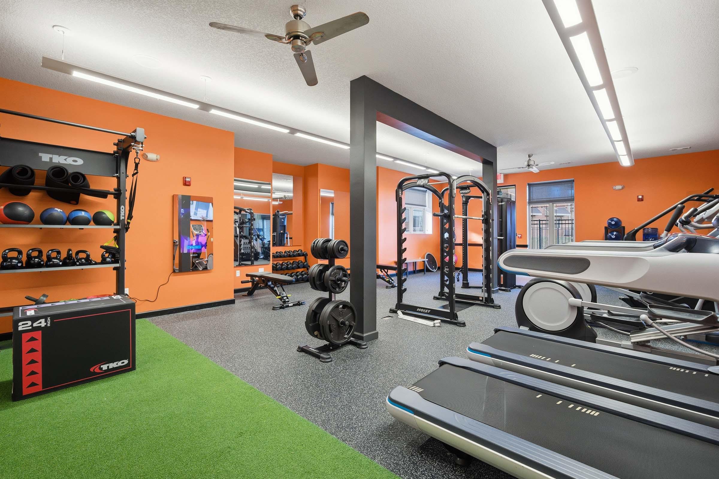 A modern gym interior featuring various fitness equipment, including weightlifting racks, dumbbells, exercise machines, and treadmills. The walls are painted orange, and there is an area with green turf. Large mirrors reflect the space, and bright overhead lights illuminate the environment.