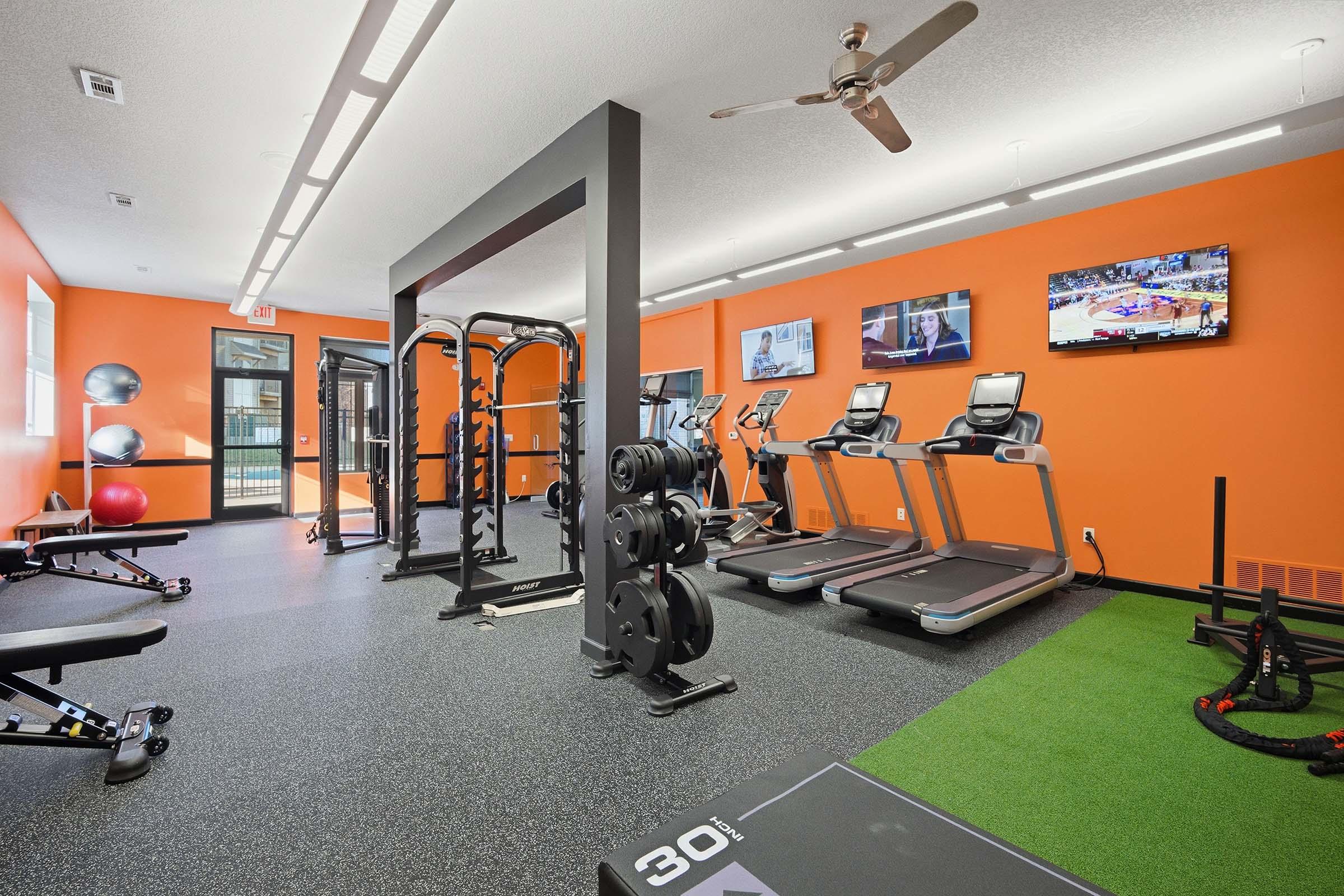 A modern gym with orange walls featuring various fitness equipment, including treadmills, a weight rack with dumbbells, exercise benches, and fitness balls. Two wall-mounted TVs display sports programs. The floor is covered in rubberized flooring and artificial turf for workout areas.