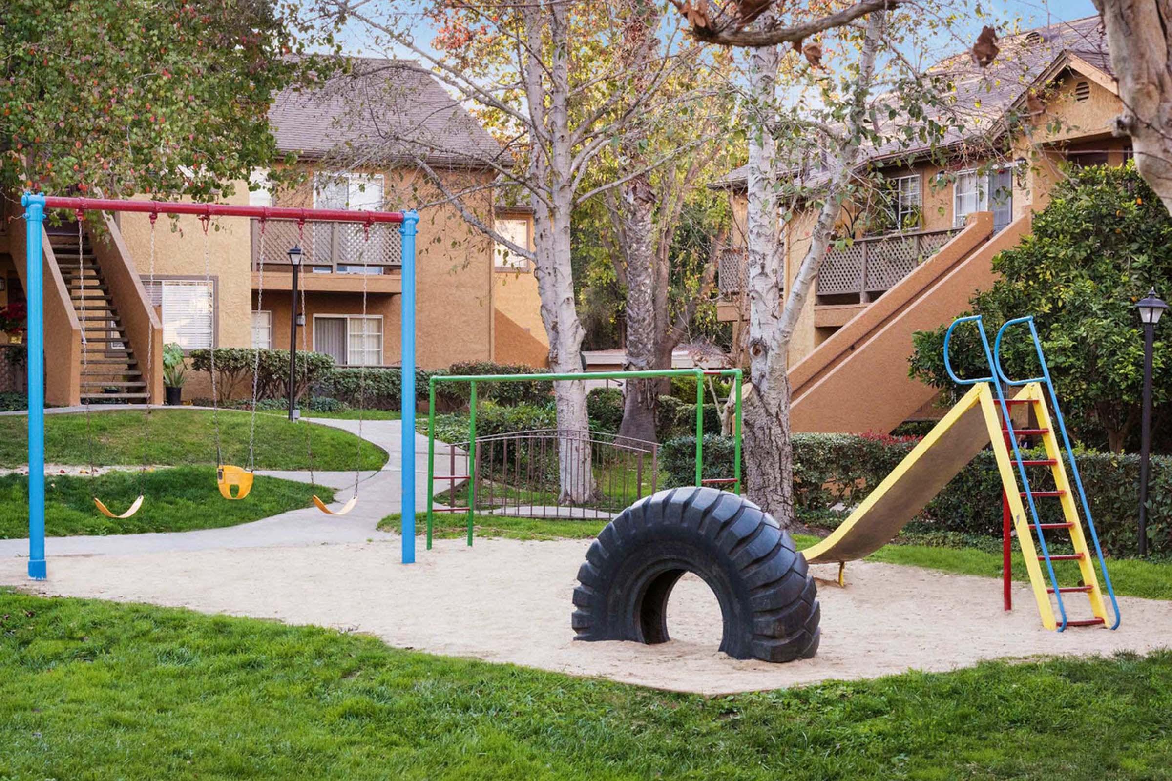 A playground featuring a tire swing, a climbing slide, and a set of swings. The area is surrounded by grass and trees, with nearby residential buildings in the background. The playground has a sandy surface, providing a playful atmosphere for children.