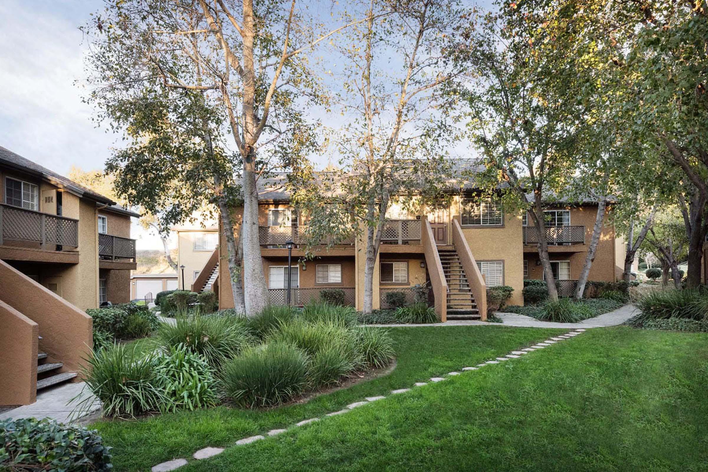 A well-maintained apartment complex surrounded by greenery, featuring multiple buildings with balconies. There are manicured lawns, trees, and a stone pathway leading through the grass, creating a serene outdoor space. The setting appears inviting and peaceful, perfect for residential living.