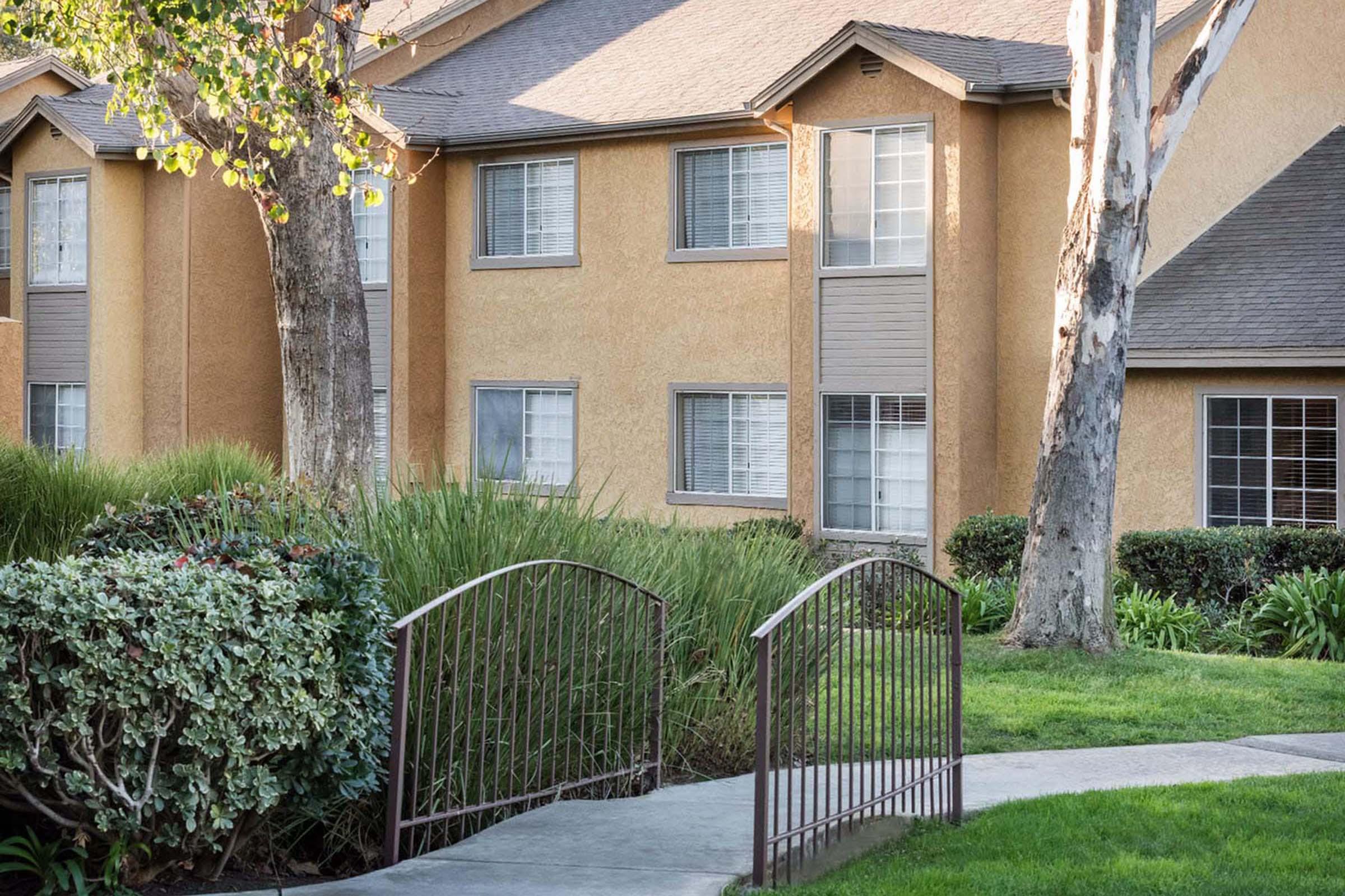A well-maintained exterior of a multi-level residential building with large windows, surrounded by lush greenery and shrubs. A curving pathway leads to a decorative metal bridge over a small area of grass. The building features a warm color palette and is framed by trees.