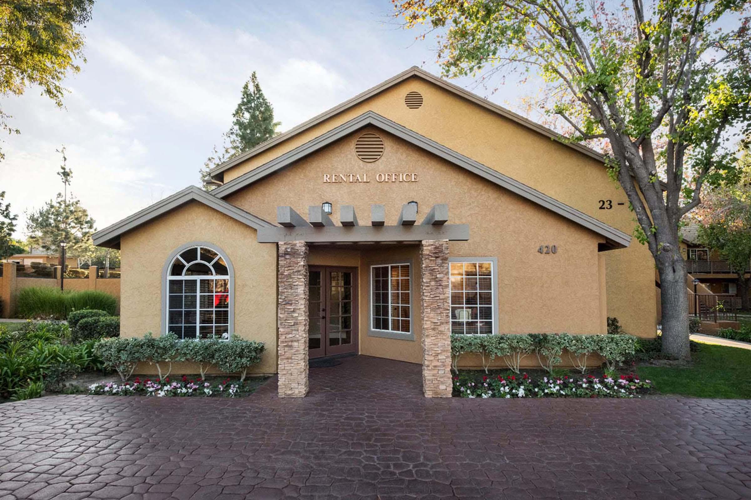 A well-maintained rental office building with a yellow exterior, stone accents, and decorative flower beds. The entrance features large windows and a decorative overhang, with the words "RENTAL OFFICE" prominently displayed above the door. Surrounding trees and landscaping enhance the welcoming atmosphere.