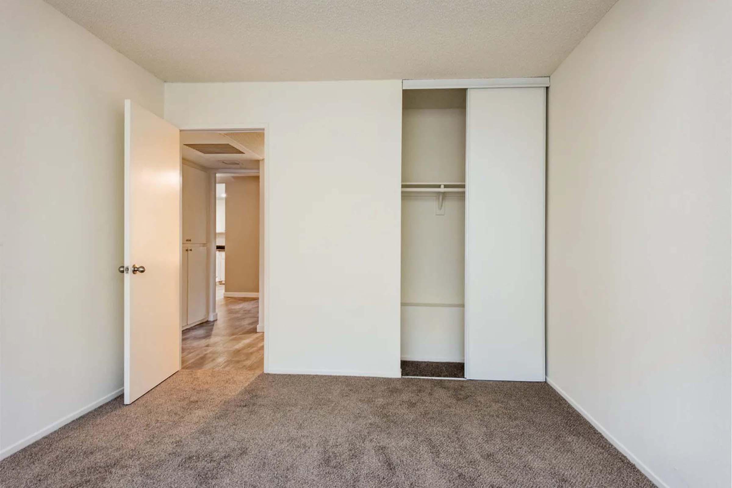 A spacious, empty bedroom featuring light-colored walls, a beige carpeted floor, and a closet with sliding doors. The room has a doorway leading to another space, enhancing its openness and potential for customization. Natural light may enter through the adjoining room.