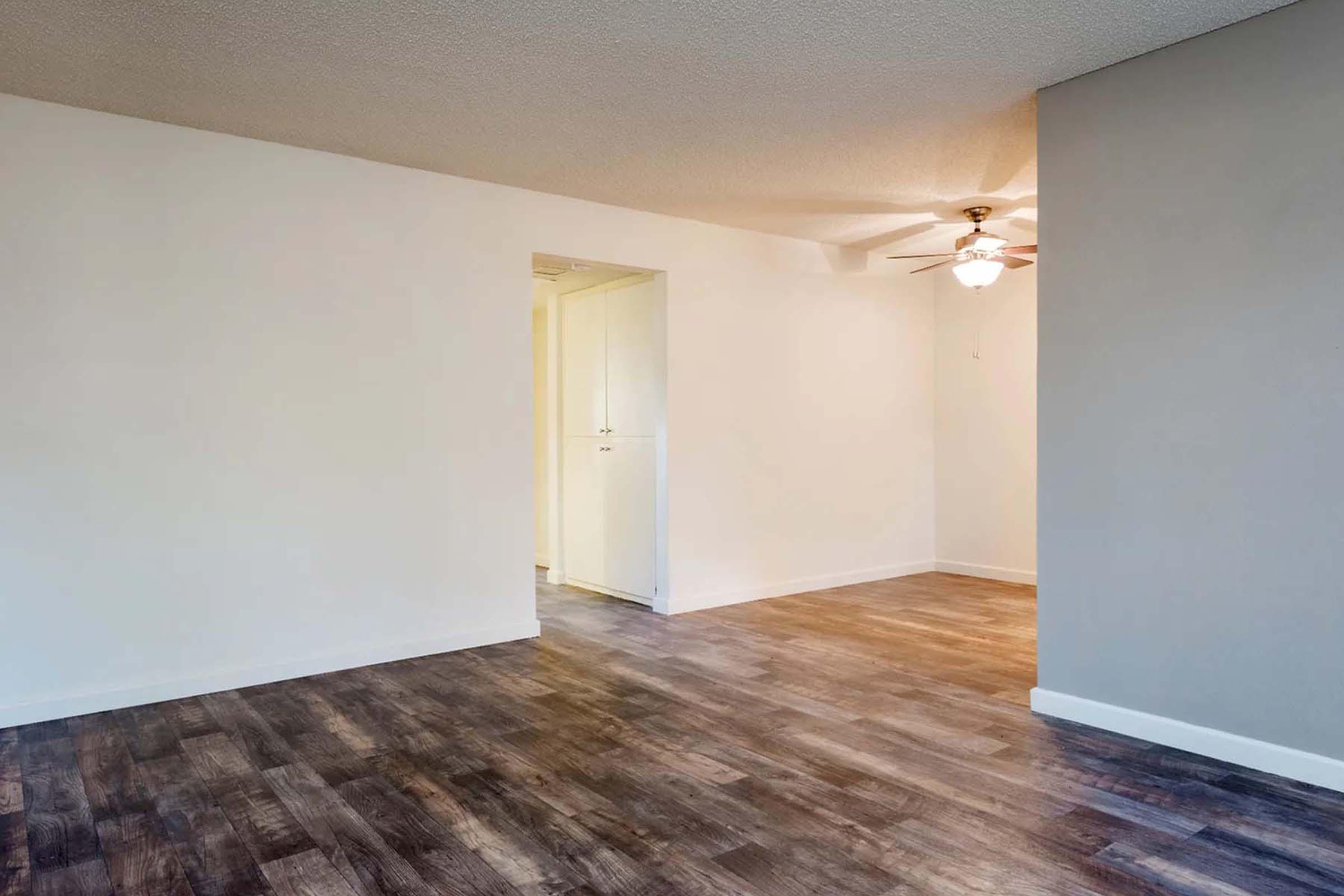 A spacious, empty room with light wood flooring and white walls. A door is visible on the left leading to another area, while a ceiling fan is mounted in the upper right corner, providing a neutral and bright atmosphere. The overall space offers potential for interior design and personalization.