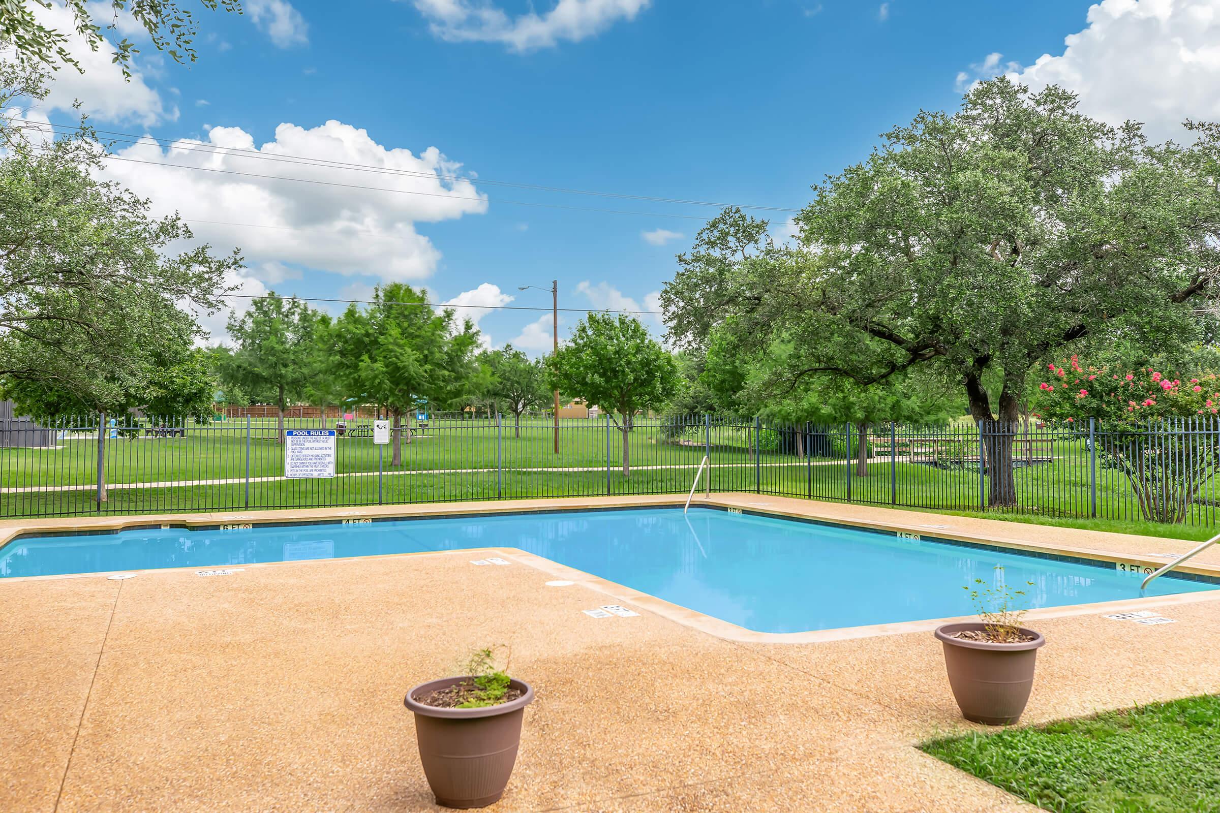 A tranquil outdoor scene featuring a clear blue swimming pool surrounded by a spacious green lawn and trees. The pool area is fenced, with potted plants on the deck. In the background, there's a grassy field with additional trees and a blue sky filled with fluffy white clouds.