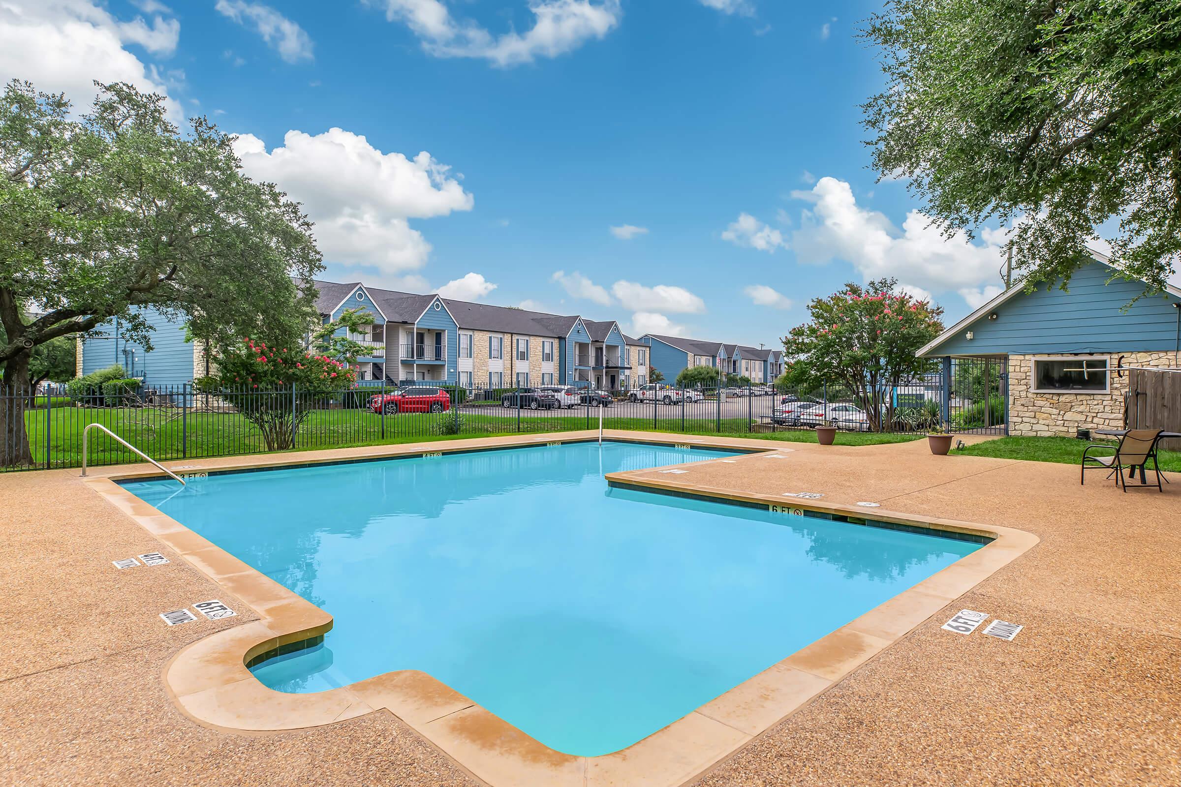 A clear, inviting swimming pool surrounded by concrete, with a gentle slope leading into the water. Lush green trees provide shade nearby, and in the background, there are residential buildings and parked cars under a bright blue sky with fluffy white clouds.