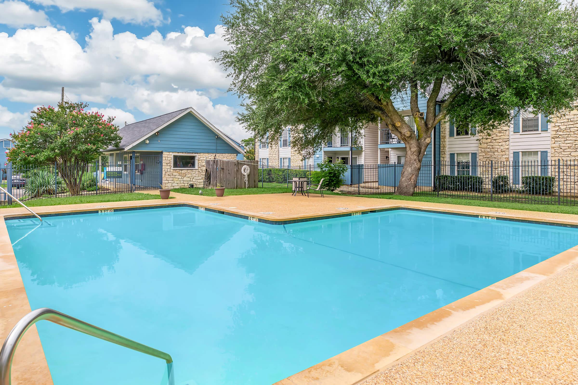 A clear blue swimming pool surrounded by green grass and trees, with a few lounge chairs nearby. In the background, there are residential buildings with stone and wooden exteriors under a partly cloudy sky. The pool area is enclosed by a black fence.