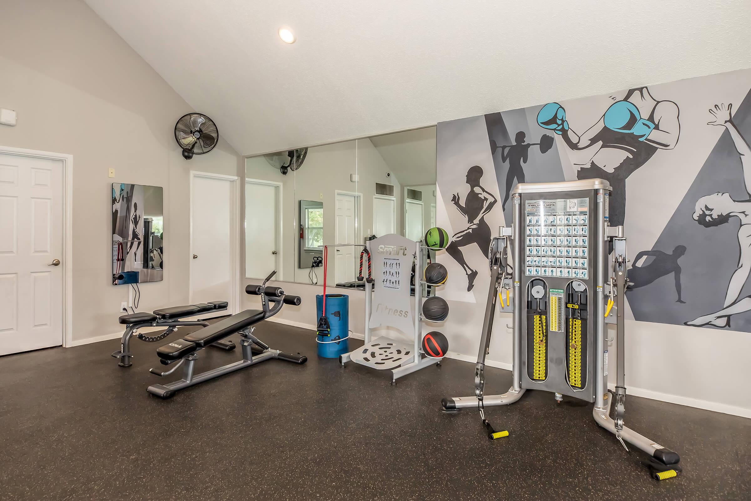 A well-equipped gym space featuring exercise benches, a multi-functional weight machine, a wall with fitness-themed artwork, large mirrors, and a fan. The flooring is dark, creating a modern and clean workout environment.
