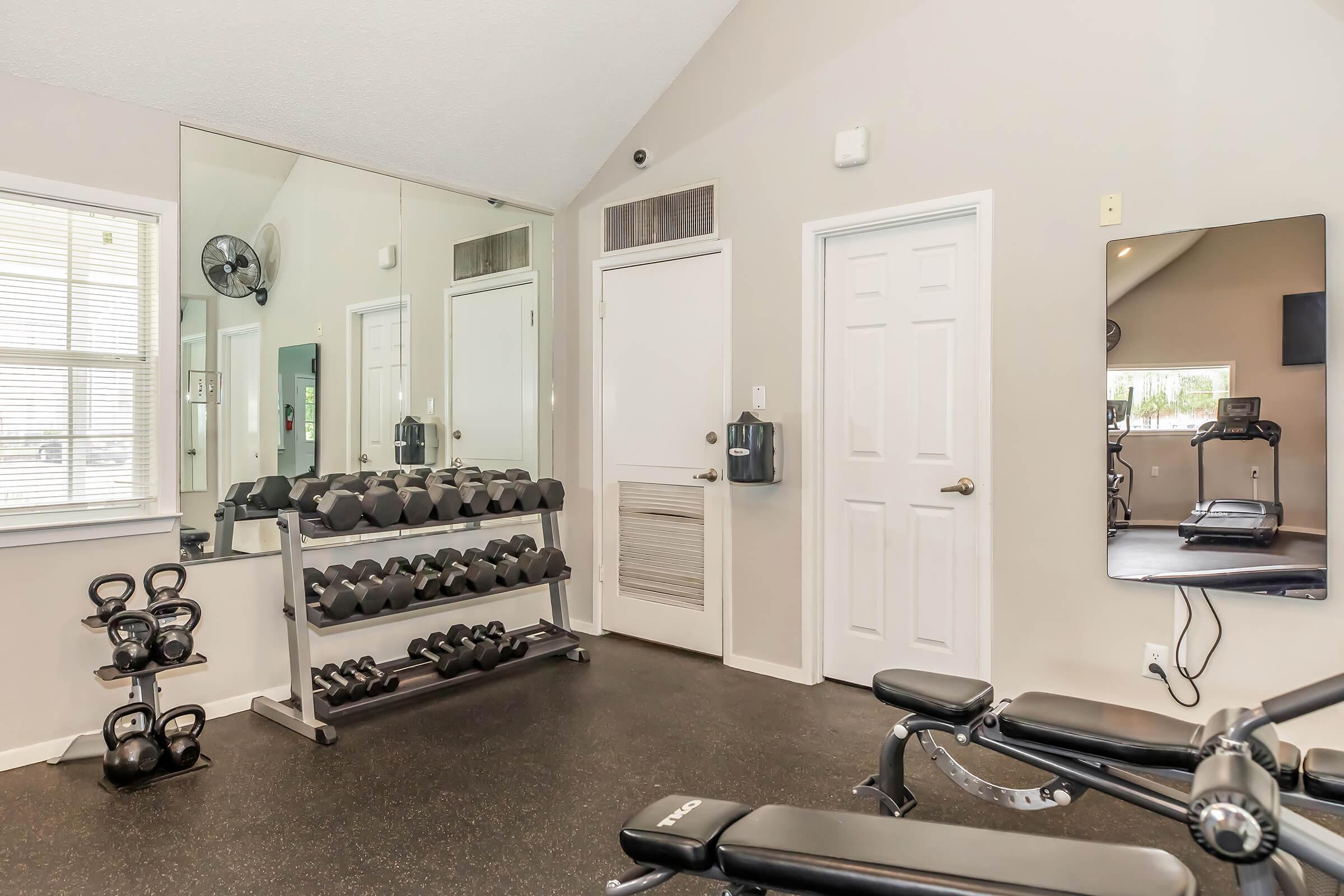 A well-lit home gym area featuring a rack of dumbbells, a bench press, and a large wall mirror. There are also kettlebells on the floor and a few exercise machines visible in the background. The walls are neutral in color, creating a clean and inviting workout space.