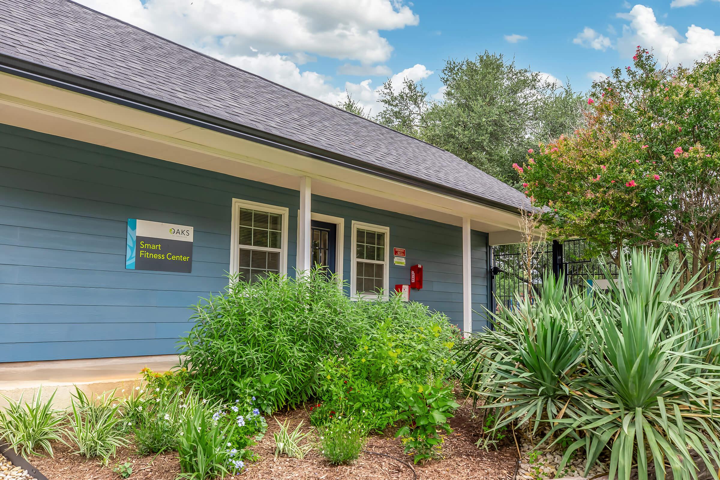 Exterior view of the Smart Fitness Center, featuring a blue building with a welcoming entrance. Surrounding the area are decorative plants and shrubs, with a clear sky above. A sign on the building indicates its name, contributing to a friendly and inviting atmosphere.