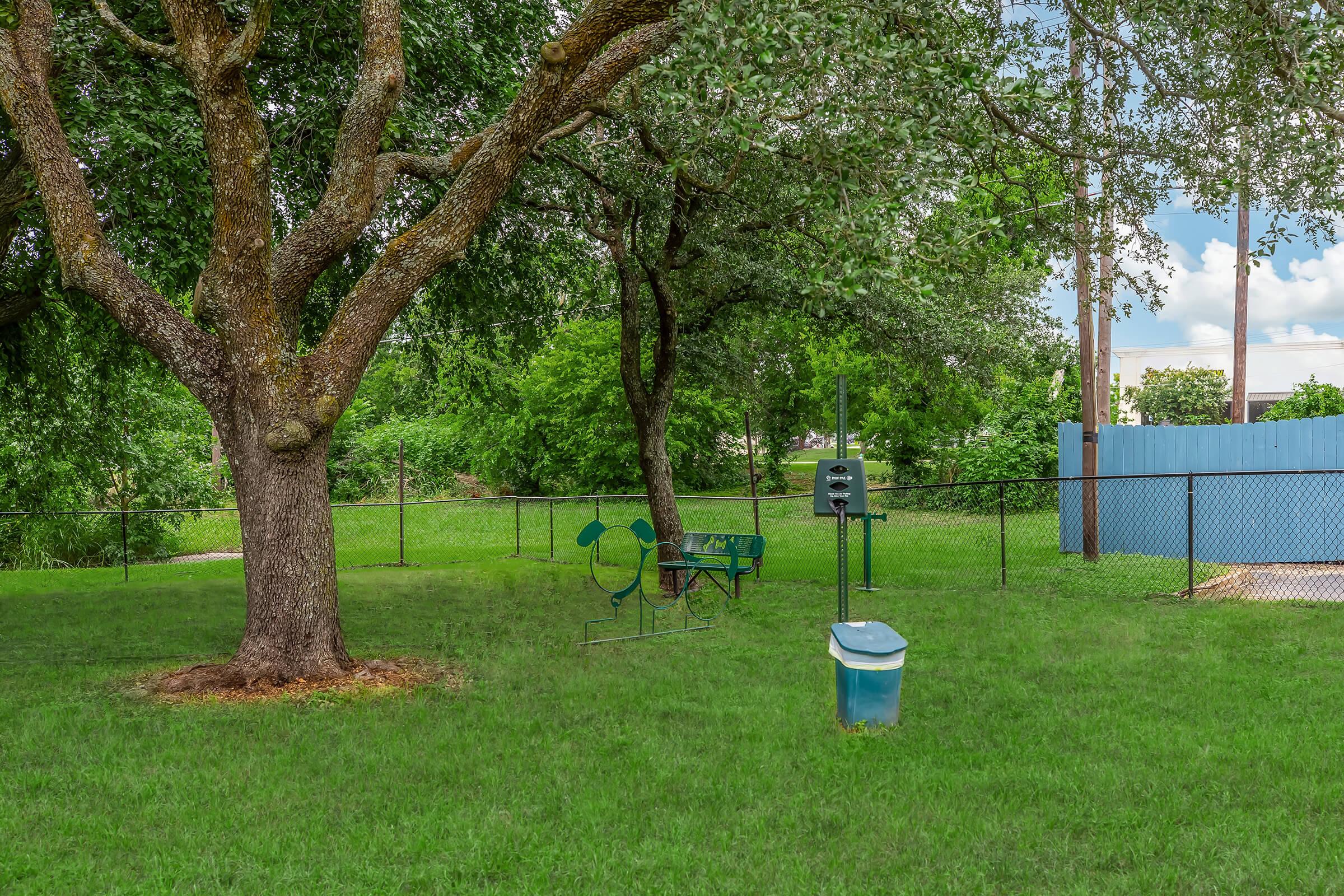 A grassy park area featuring a large tree, a green bench, and a small waste bin. In the background, there is a fenced area and a blue structure, with trees and a cloudy sky visible. The setting suggests a peaceful outdoor space suitable for relaxation or leisure activities.