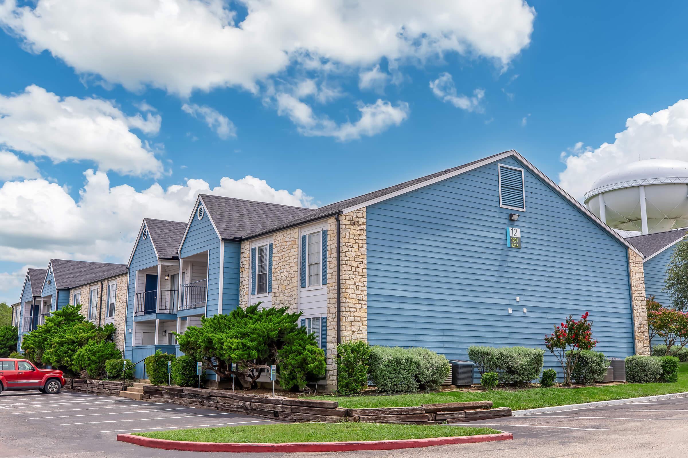 A residential building with a blue exterior, featuring two stories and balconies. The landscaping includes greenery and shrubbery in front. A red car is parked nearby, and a water tower is visible in the background against a blue sky with fluffy white clouds.
