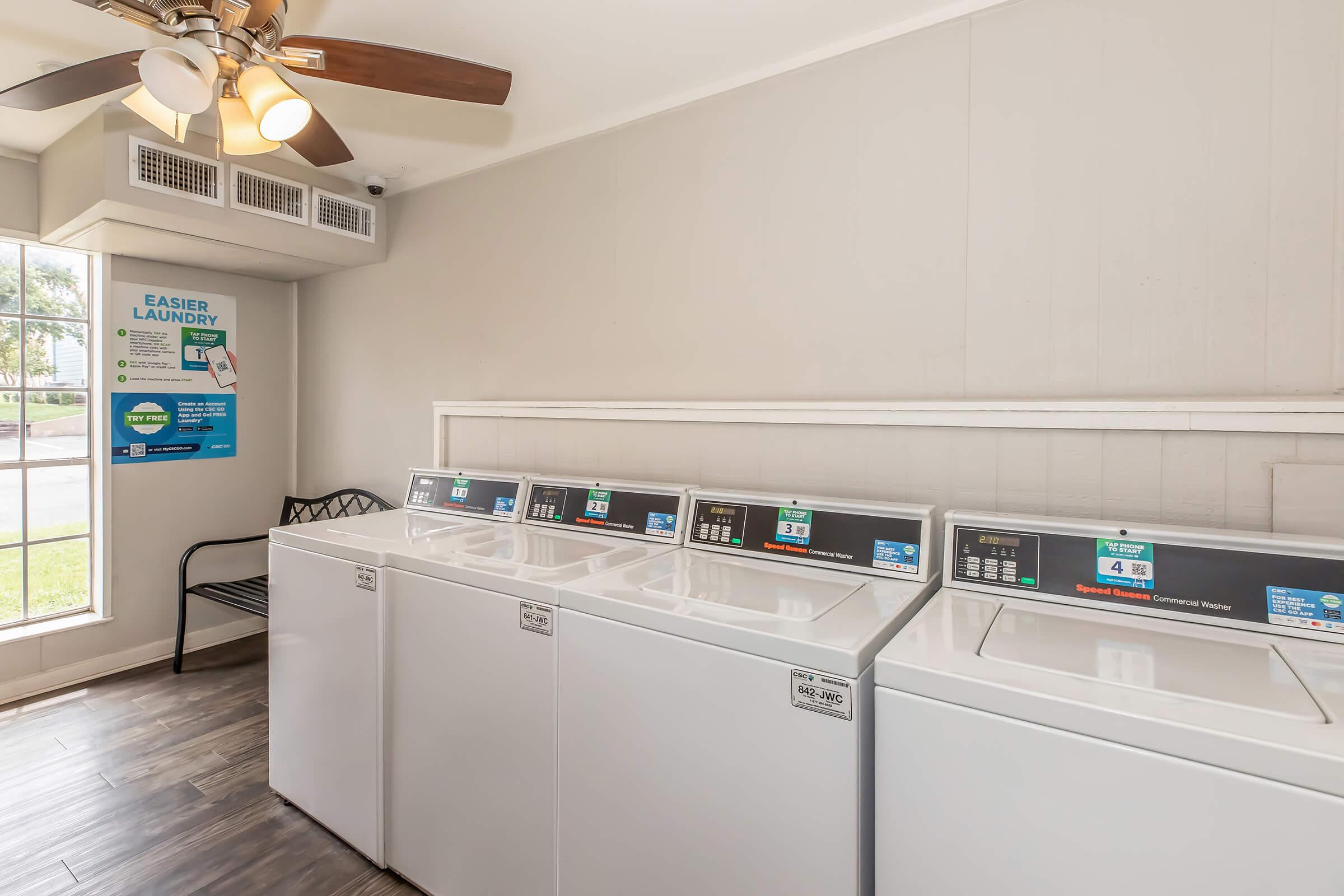 A clean and modern laundry room with three white washing machines arranged side by side. A black chair is against the wall, and a ceiling fan is visible above. A wall poster promotes easy laundry services, while natural light filters in through the windows, creating a bright atmosphere.