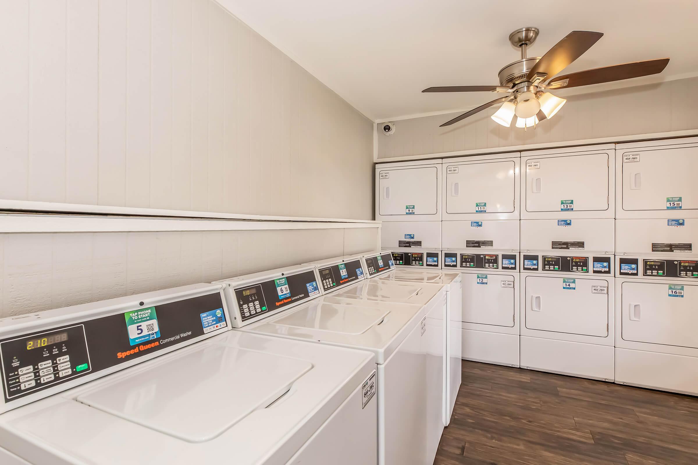 A laundry room featuring several white washing machines and dryers lined up against the wall. The machines have digital displays and buttons. There's a ceiling fan overhead, and the room has light-colored walls and dark wooden flooring.