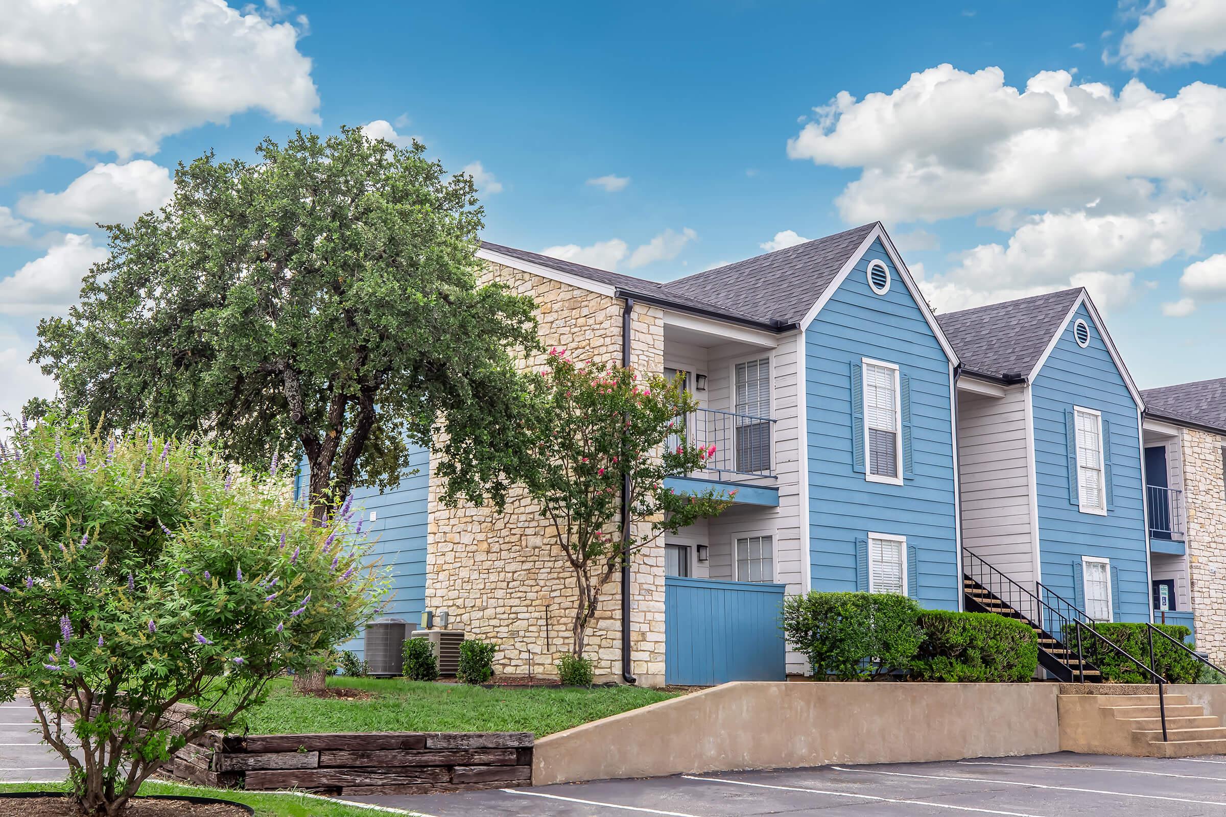 A residential apartment complex featuring two buildings, one with stone and the other with blue siding. Lush greenery, including a large tree and flowering plants, surround the area. The sky is partly cloudy, adding a pleasant ambiance to the scene.