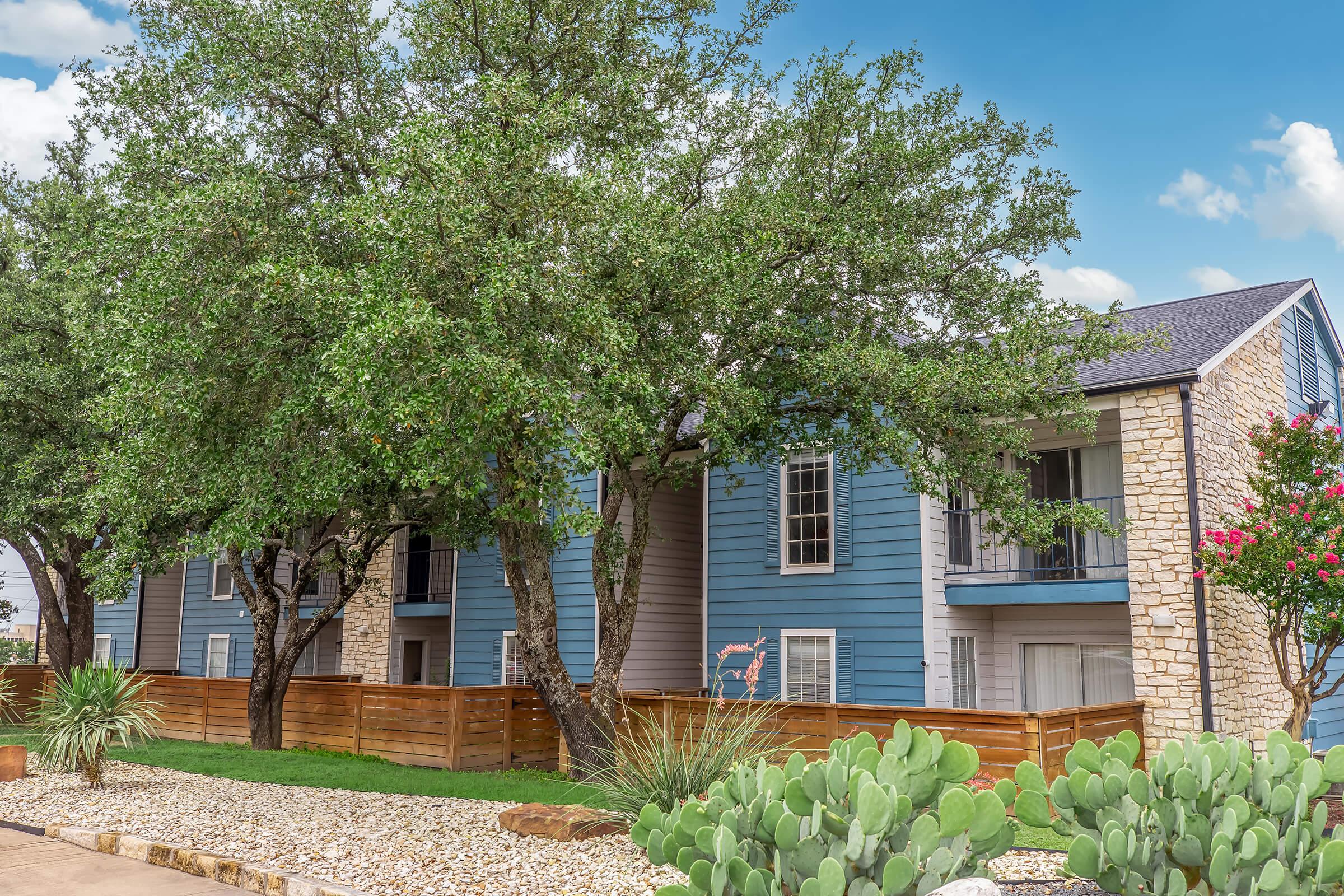 A two-story blue apartment building with stone accents, surrounded by leafy trees and shrubs. The landscape features gravel and cacti, with wooden fencing creating a cozy outdoor environment. The sky is partly cloudy, adding to the serene setting.