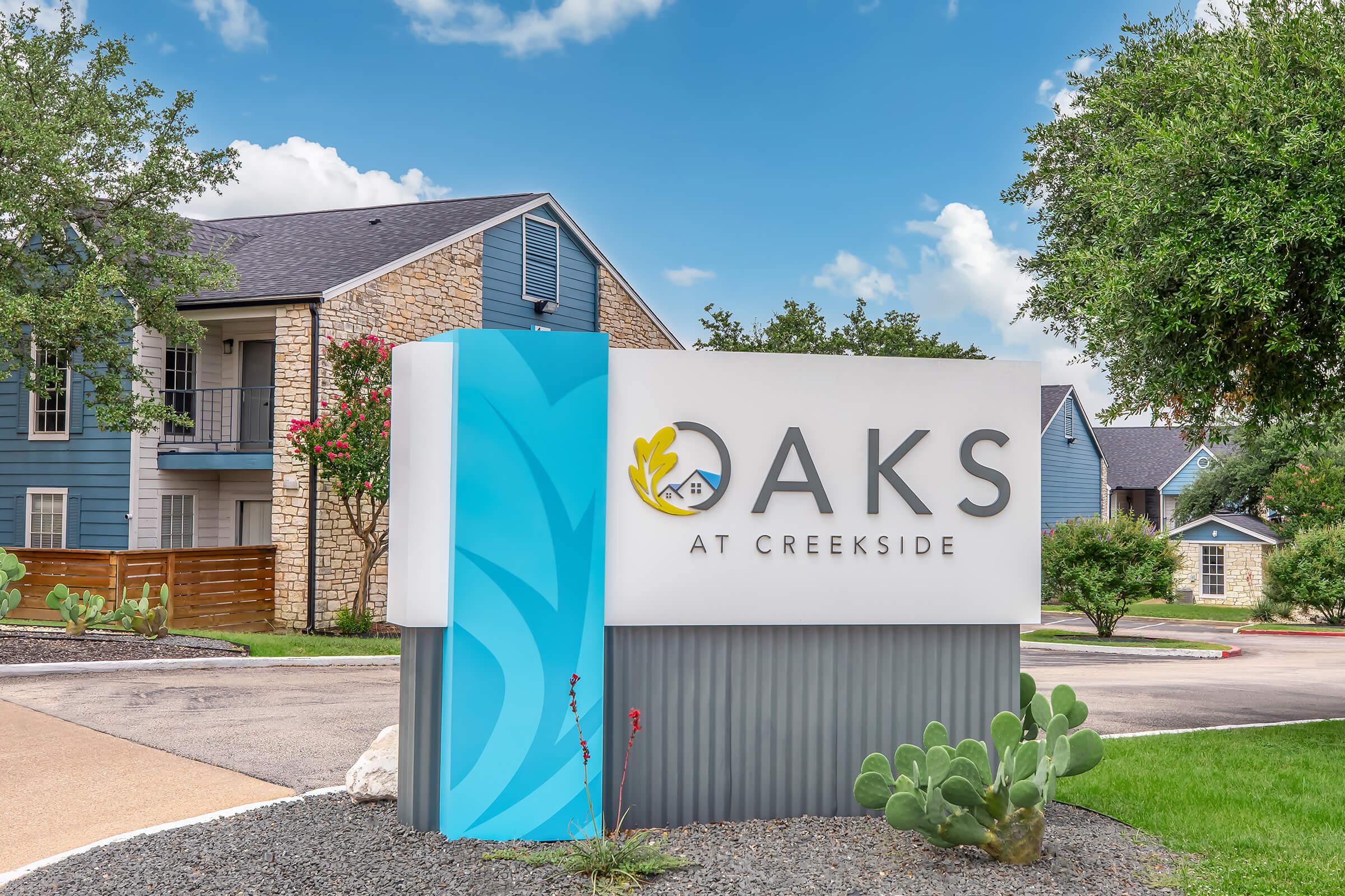 Sign for "Oaks at Creekside" featuring a modern design with a blue and white color scheme. Surrounding landscape includes greenery and cacti, indicative of a residential community setting. Bright, sunny sky with scattered clouds in the background.