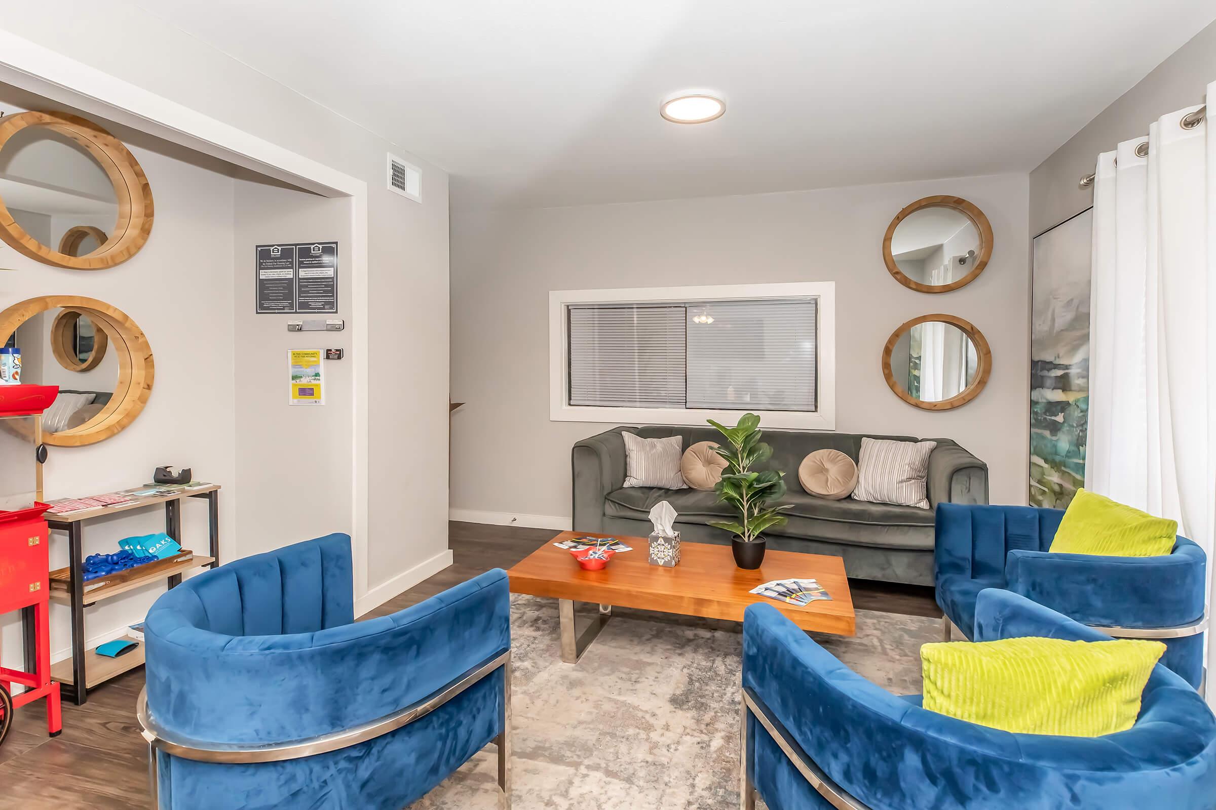 Cozy living area featuring a green sofa with decorative pillows, flanked by four blue accent chairs. A wooden coffee table sits in the center, adorned with a small plant and magazines. The walls are decorated with round mirrors and artwork, creating a welcoming atmosphere. Natural light filters through sheer curtains.