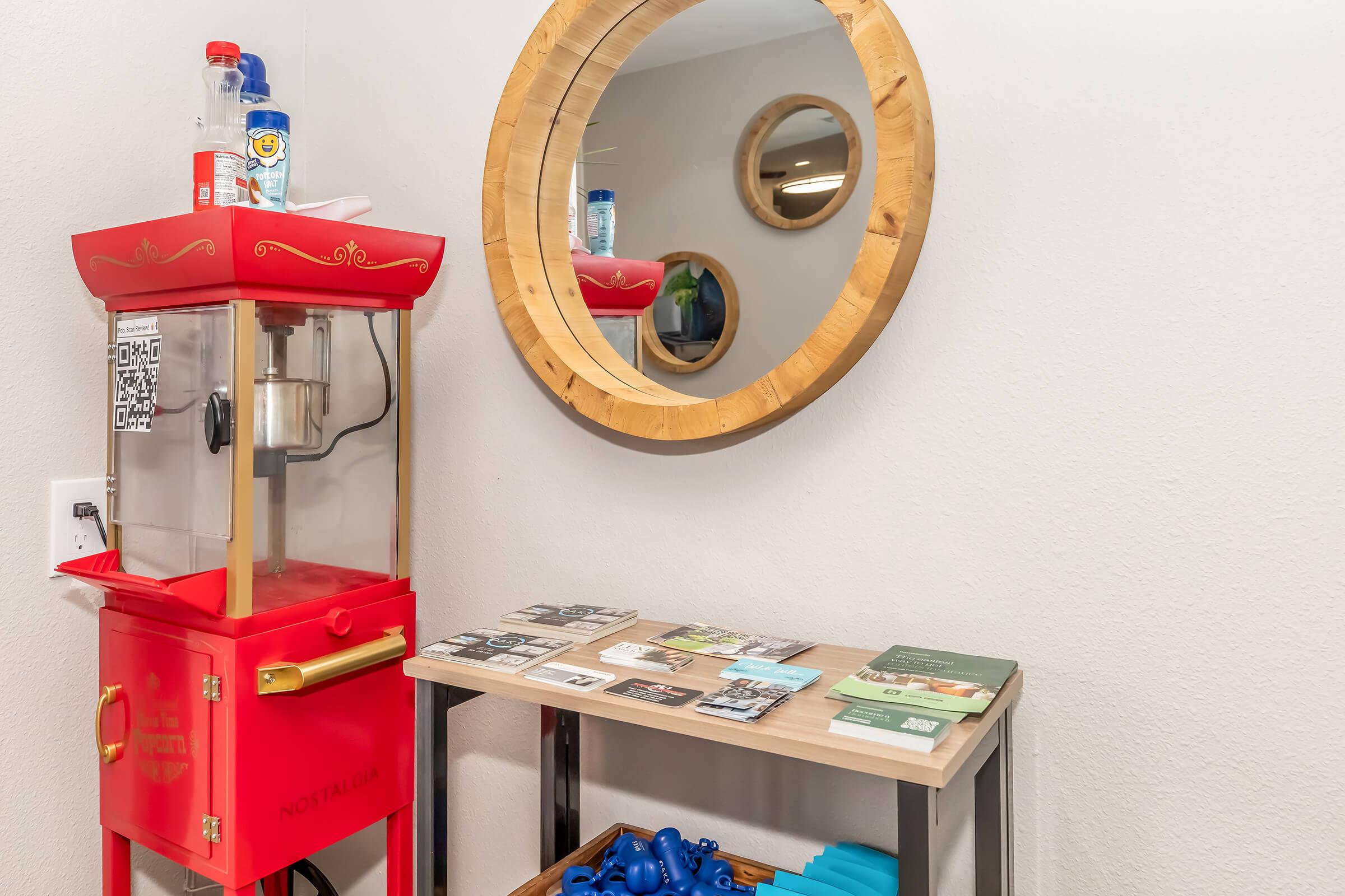 A vintage popcorn machine in red, positioned next to a round wooden mirror on a wall. Below the mirror, there's a wooden table displaying various brochures and a container filled with blue fitness accessories. The background features a simple, light-colored wall.
