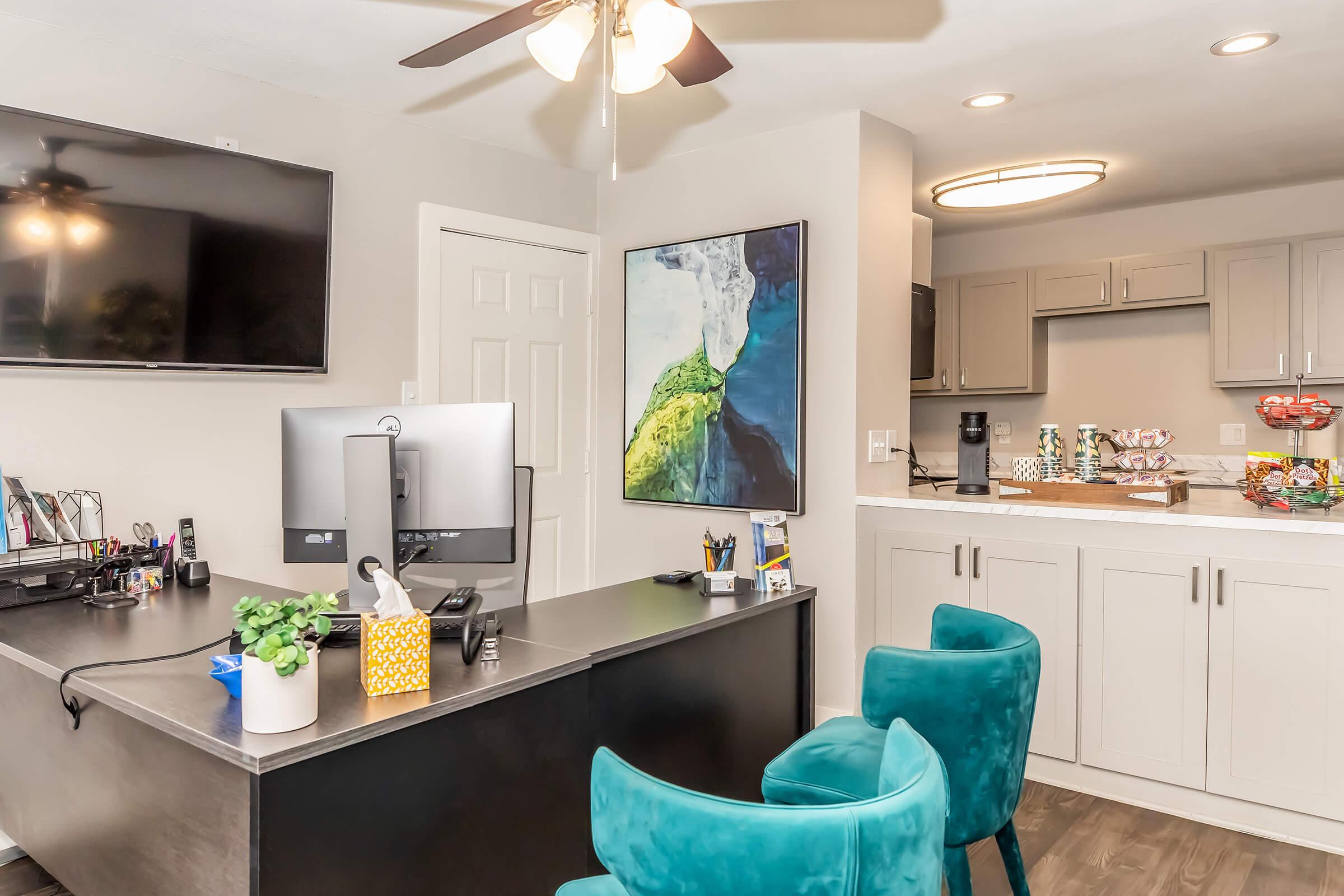 An office space featuring a dark wooden desk with a computer monitor, surrounded by teal chairs. There’s a wall-mounted TV and an abstract painting depicting green and blue colors. The room includes a kitchenette area with cabinets and snacks on the counter, along with a potted plant on the desk.