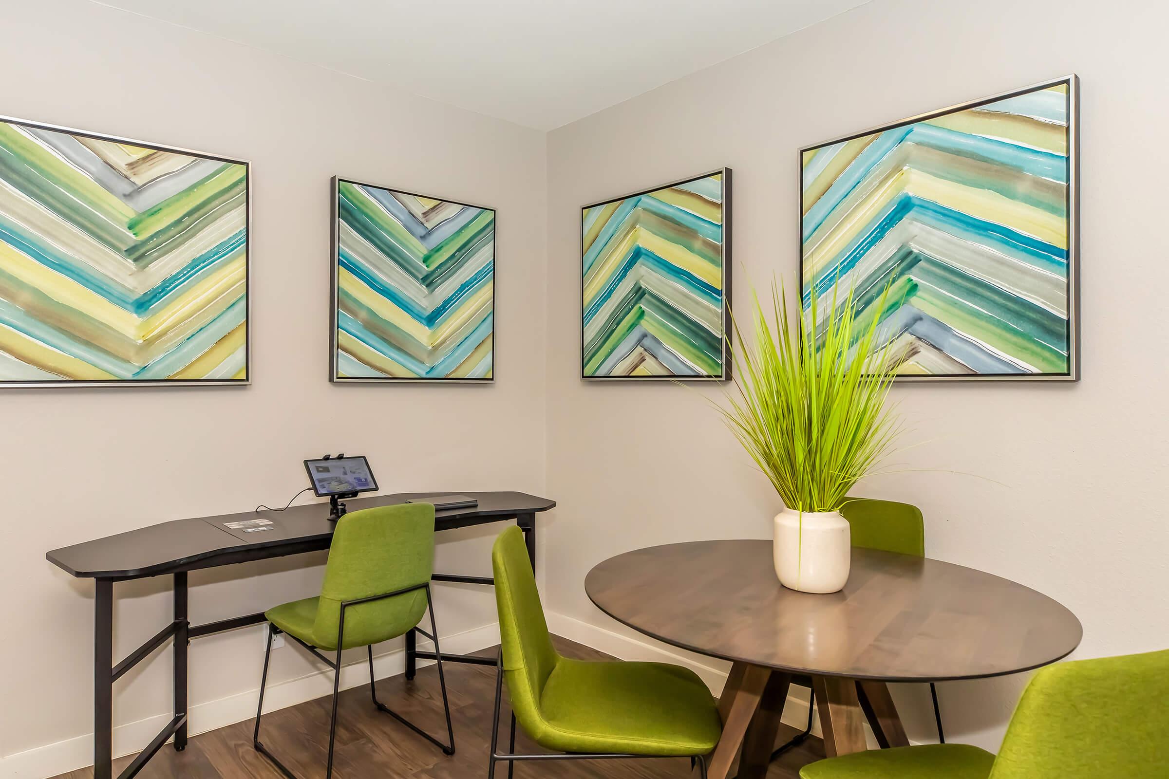 A cozy office space featuring a round wooden table with green chairs, a simple desk with a small device, and three vibrant chevron-patterned wall art pieces in green and blue tones. A potted plant adds a touch of greenery to the room, creating a welcoming atmosphere for work or study.