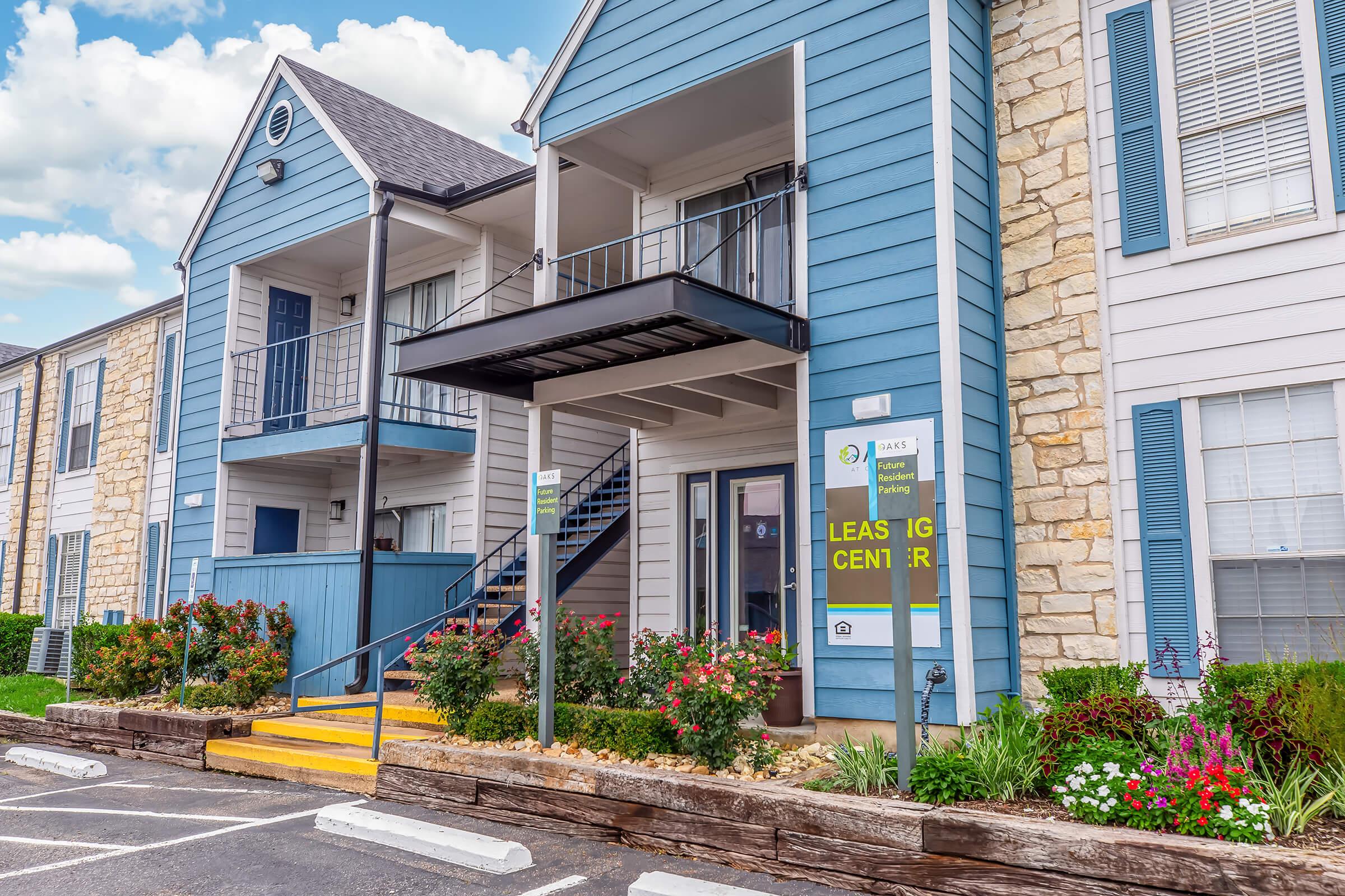 A modern apartment complex featuring a leasing center entrance. The building has blue and beige exteriors with stone accents. There are flower beds with colorful plants, steps leading to the entrance, and a sign indicating it is the leasing center. The sky is partly cloudy.