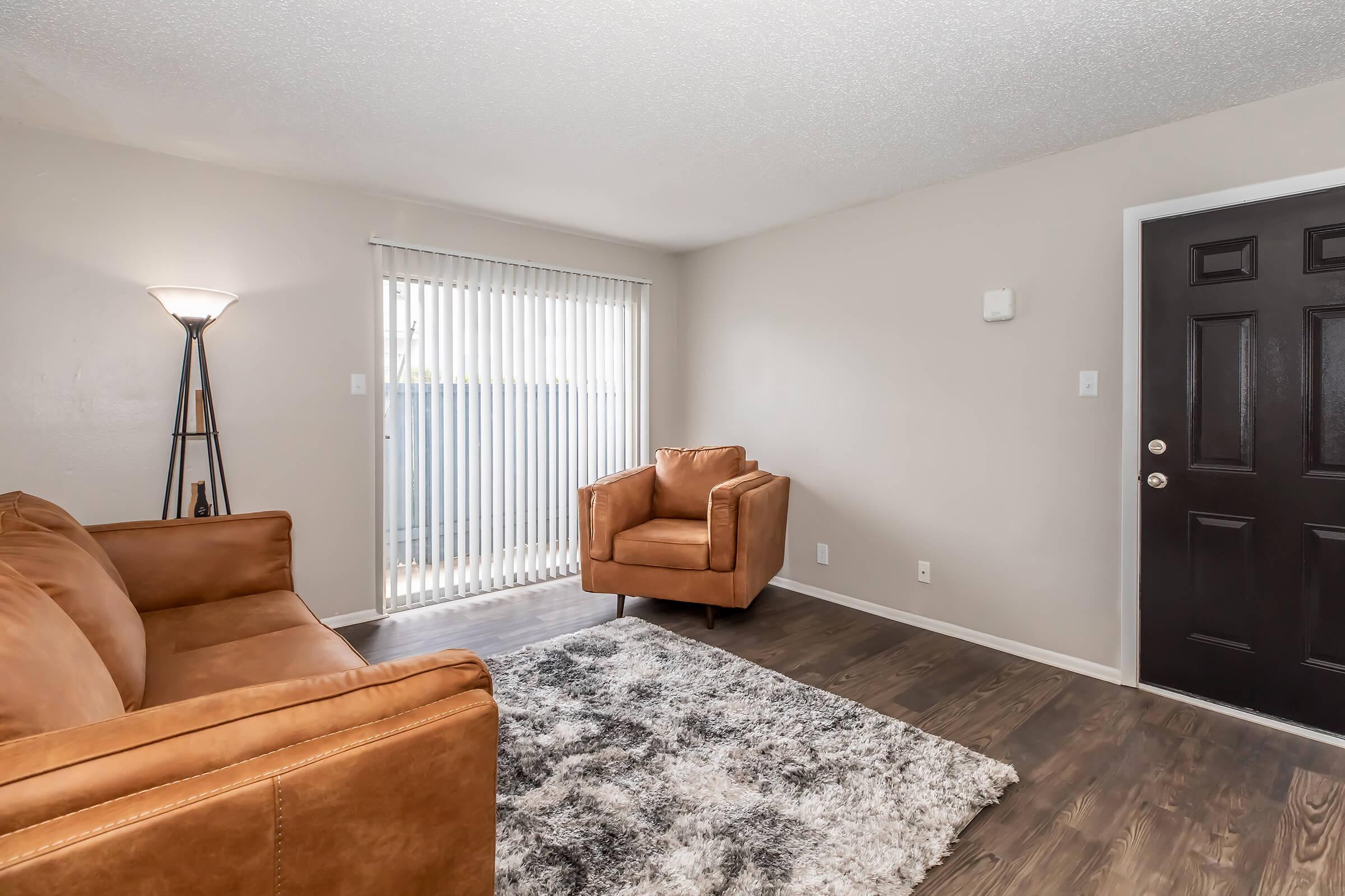 Cozy living room featuring a brown leather sofa and an armchair. There is a textured white rug on a hardwood floor. Natural light filters through vertical blinds, and a modern floor lamp provides additional lighting. A dark front door is visible on the right side of the image.