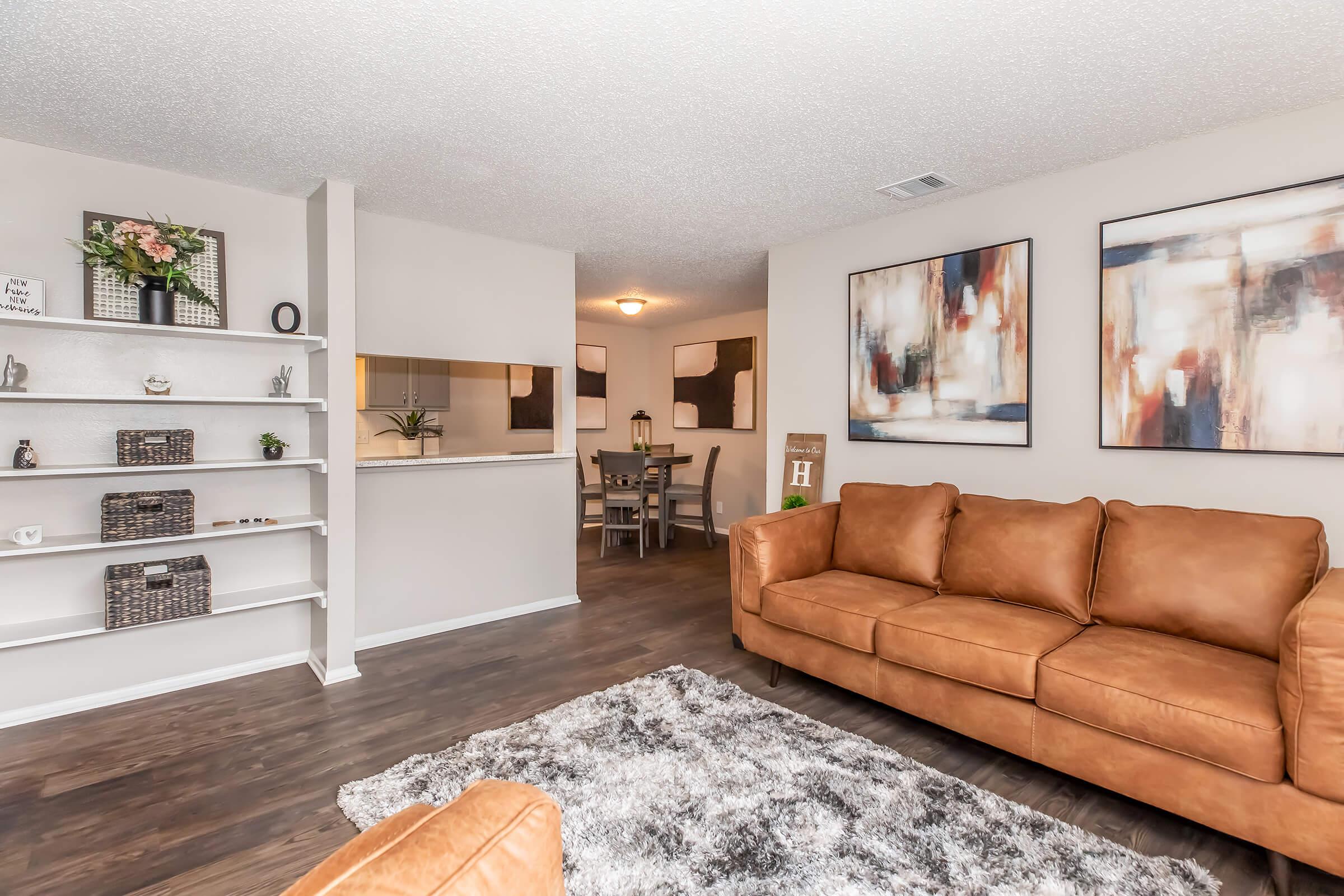 A cozy, modern living room featuring a brown leather sofa, a plush area rug, and decorative wall art. To the left, a white shelf displays decorative items, while a dining area with a table and chairs is visible in the background. The space is bright and inviting, with neutral tones and warm lighting.