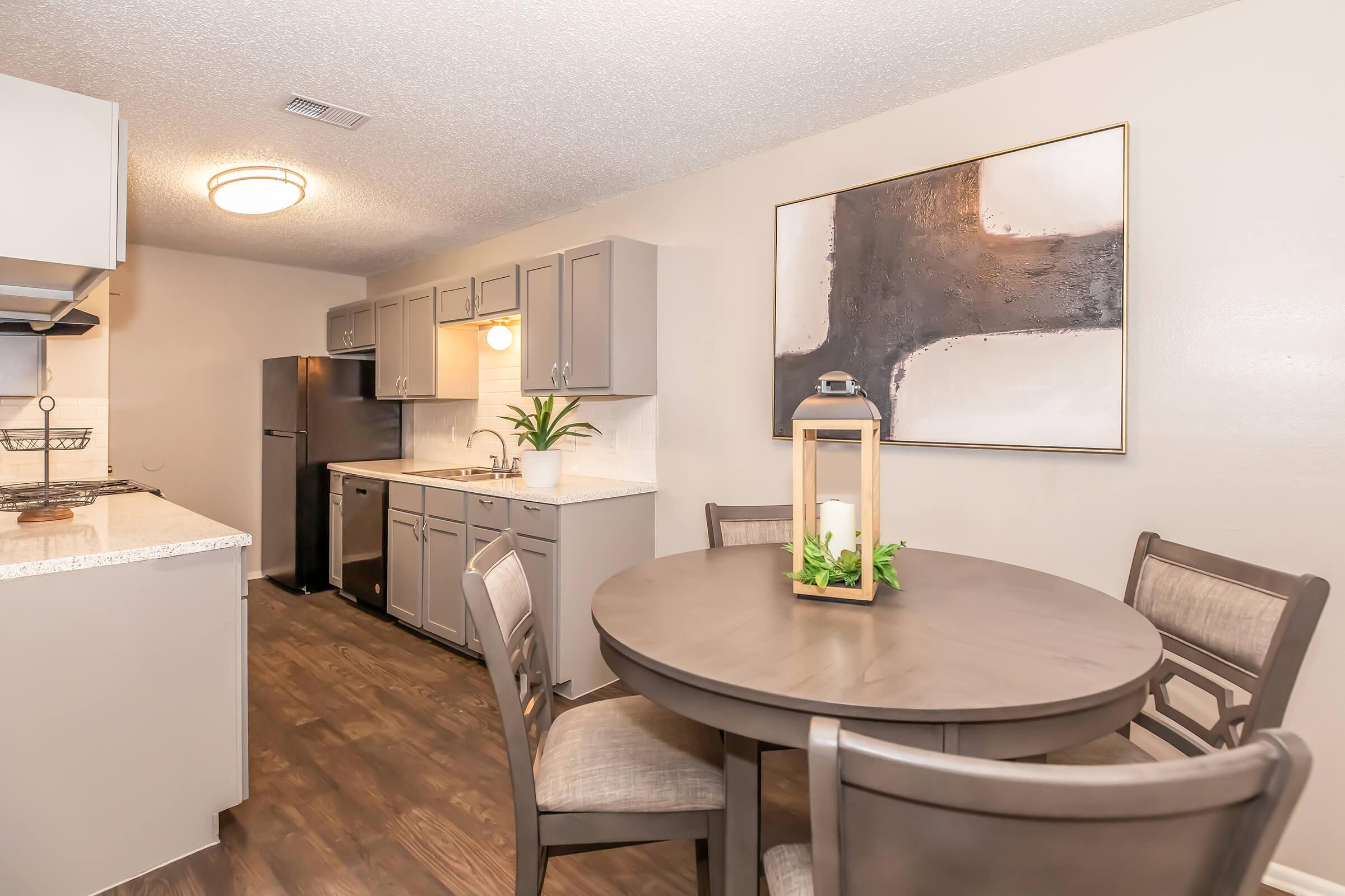 Modern kitchen and dining area featuring gray cabinets, a circular wooden table with four chairs, and a decorative lantern centerpiece. The kitchen includes a black refrigerator and a light fixture above the table, with a neutral-tone artwork on the wall. A potted plant adds a touch of greenery.