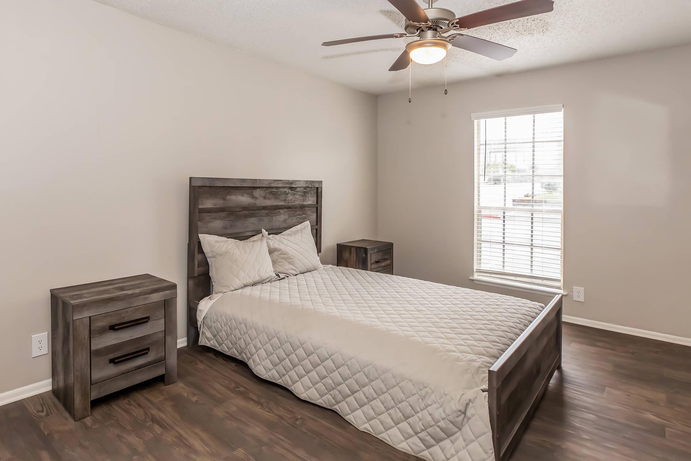 A simple bedroom featuring a wooden bed frame with a light-colored quilt, two matching nightstands on either side, and a ceiling fan. Natural light streams through a window with blinds, highlighting the neutral-colored walls and wooden flooring.