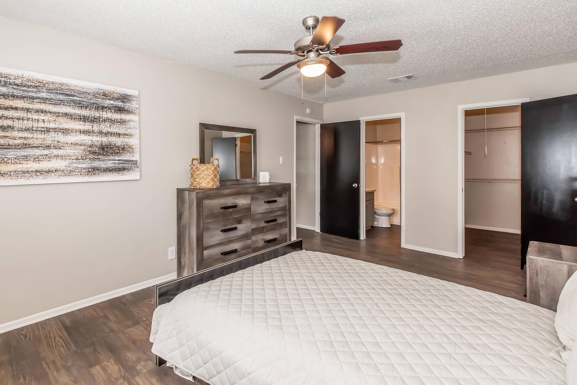 A well-decorated bedroom featuring a bed with a quilted white comforter, a dark wood dresser, and a wall-mounted mirror. The room has a ceiling fan and neutral-colored walls. Two doors lead to a bathroom and a closet area, enhancing the spacious feel of the room.