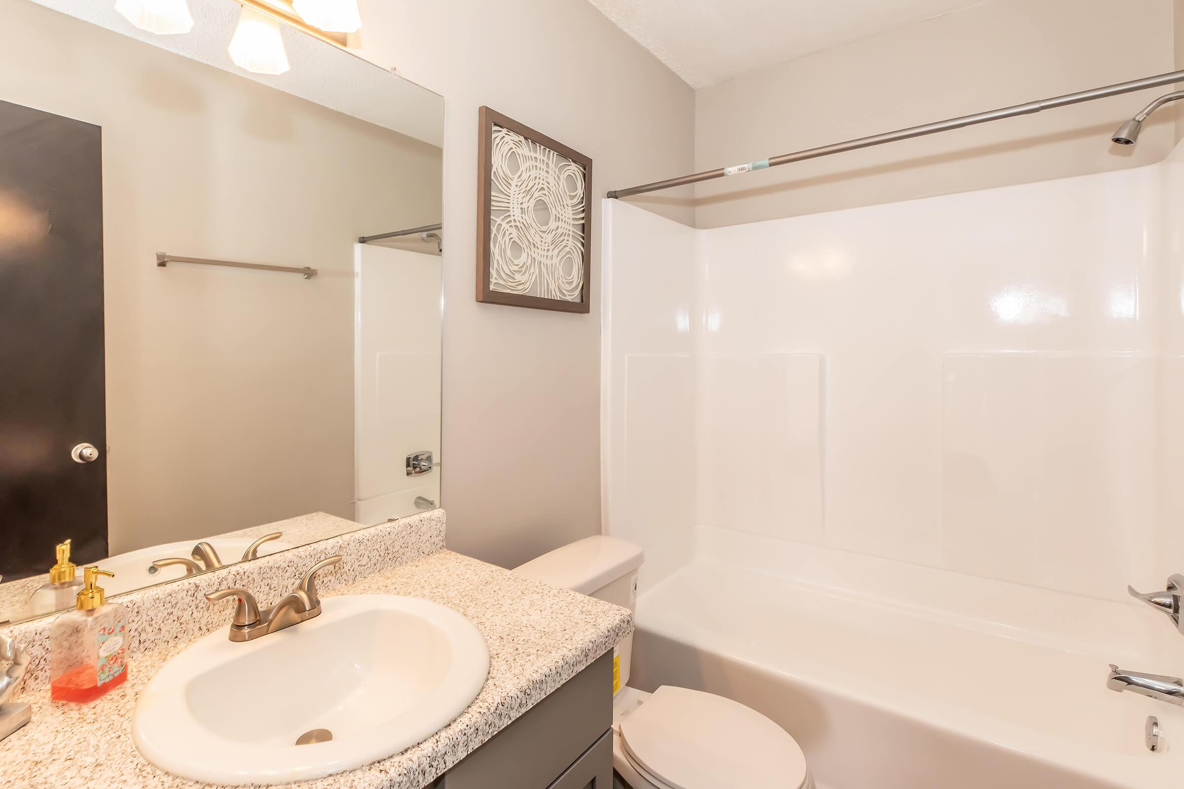 A clean and modern bathroom featuring a gray vanity with a granite countertop, a round sink, and a shower-tub combination. The walls are painted a light color, and there is a mirror above the sink. A decorative wall piece is mounted beside a towel rack, enhancing the overall aesthetic.