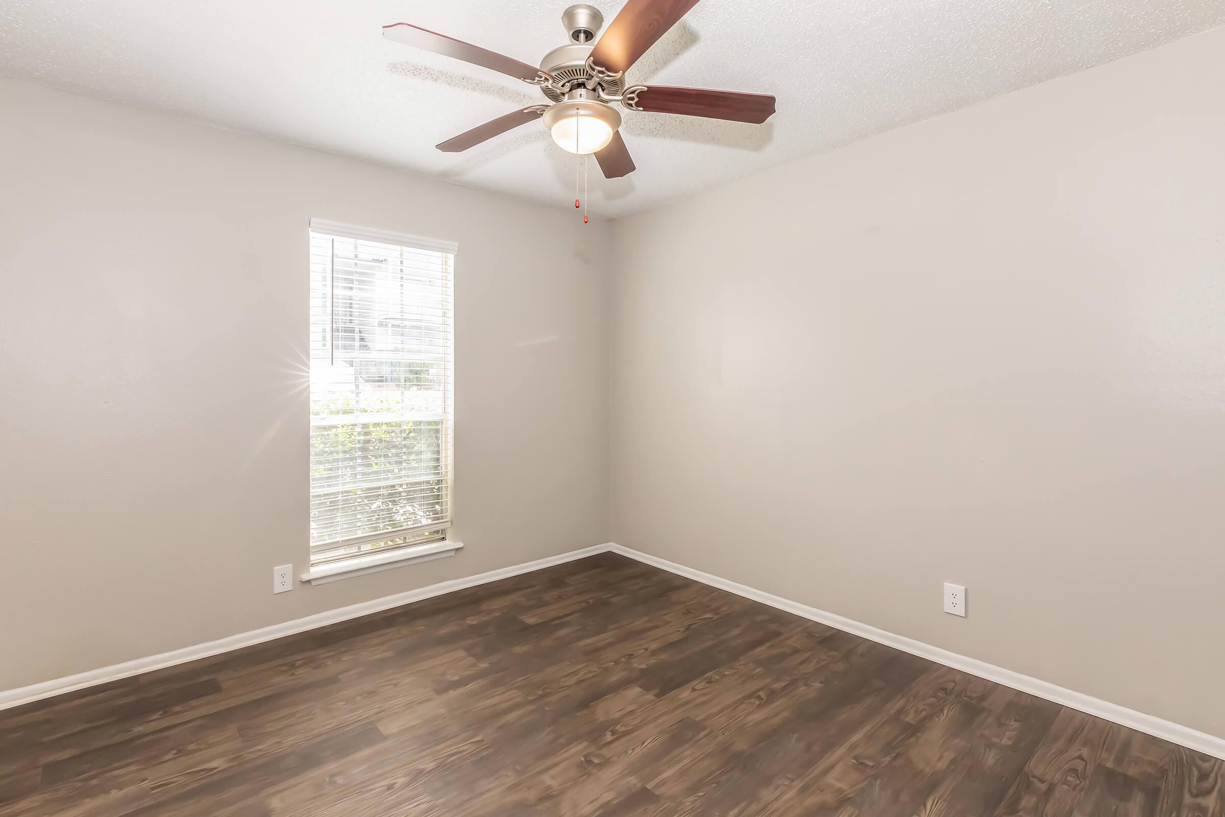 A well-lit, empty bedroom featuring a ceiling fan with wooden blades. The walls are painted a soft gray, and there is a window allowing natural light to enter. The floor is covered with dark wood laminate, creating a warm and inviting atmosphere.