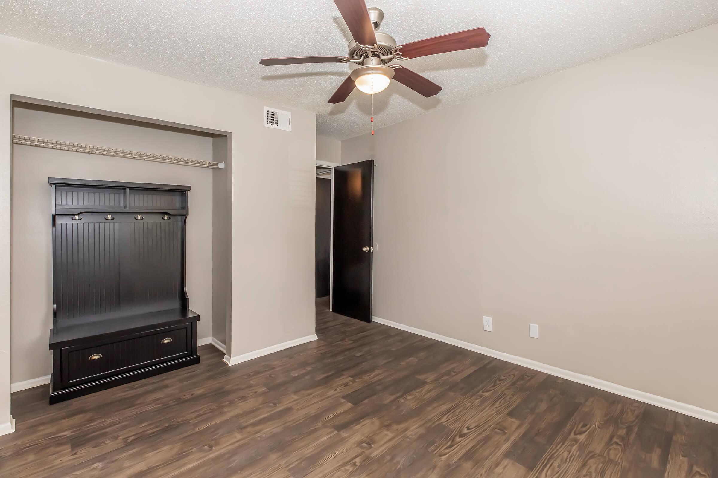 A spacious room featuring a ceiling fan, light-colored walls, and dark wood flooring. On one side, there is a black wooden coat rack with storage below, and a doorway leading to another part of the space. The overall atmosphere is bright and inviting.