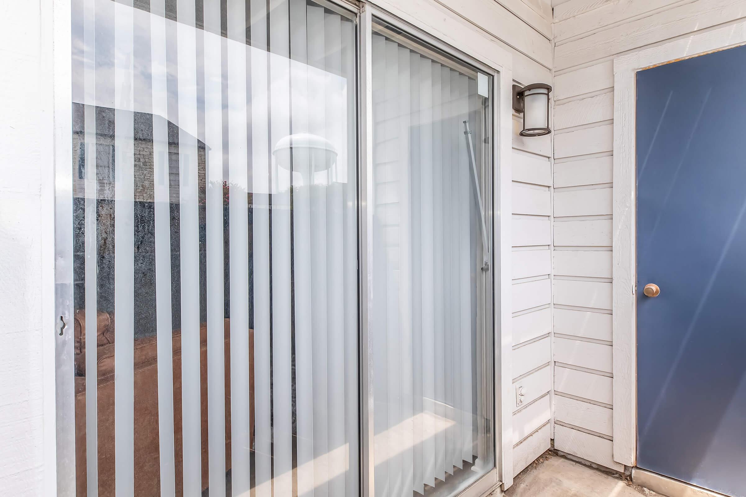 Sliding glass doors with vertical blinds, located next to a blue door. The exterior features white wooden siding, and there is a shadow of a nearby structure reflected on the glass. The area appears to be well-lit with natural light.