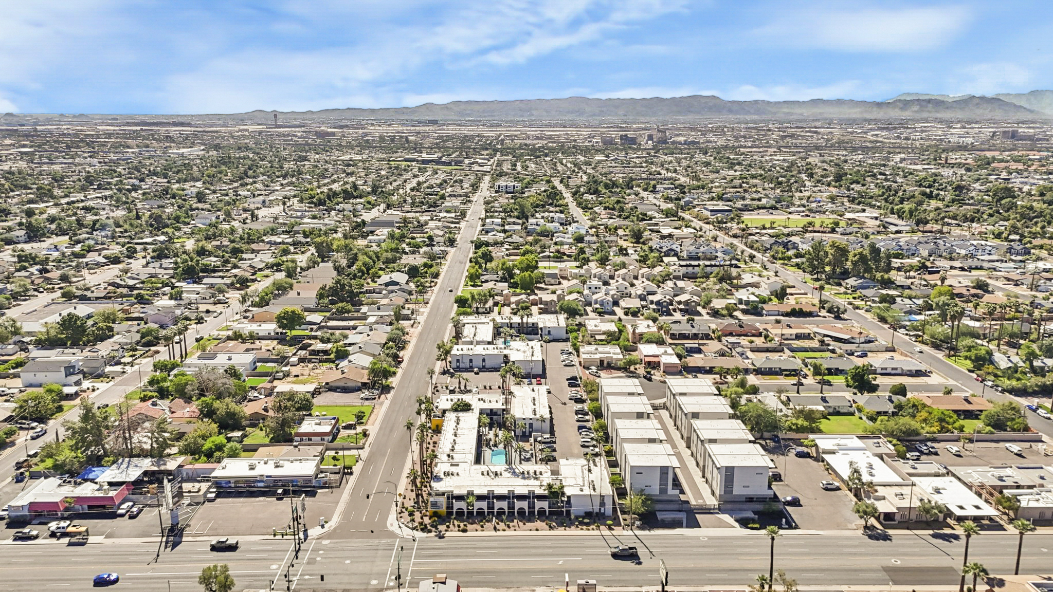 Aerial view of a suburban area featuring a mix of residential homes and commercial buildings. The landscape includes streets, greenery, and distant mountains under a clear blue sky. The scene illustrates a sprawling neighborhood layout with various structures visible.
