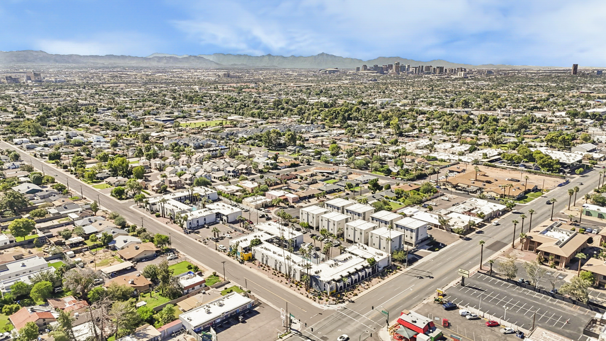 Aerial view of a suburban landscape featuring a mix of residential neighborhoods, commercial buildings, and green spaces. In the background, a city skyline with tall buildings is visible under a clear blue sky, while mountains can be seen in the distance. The image captures the contrast between urban and suburban areas.