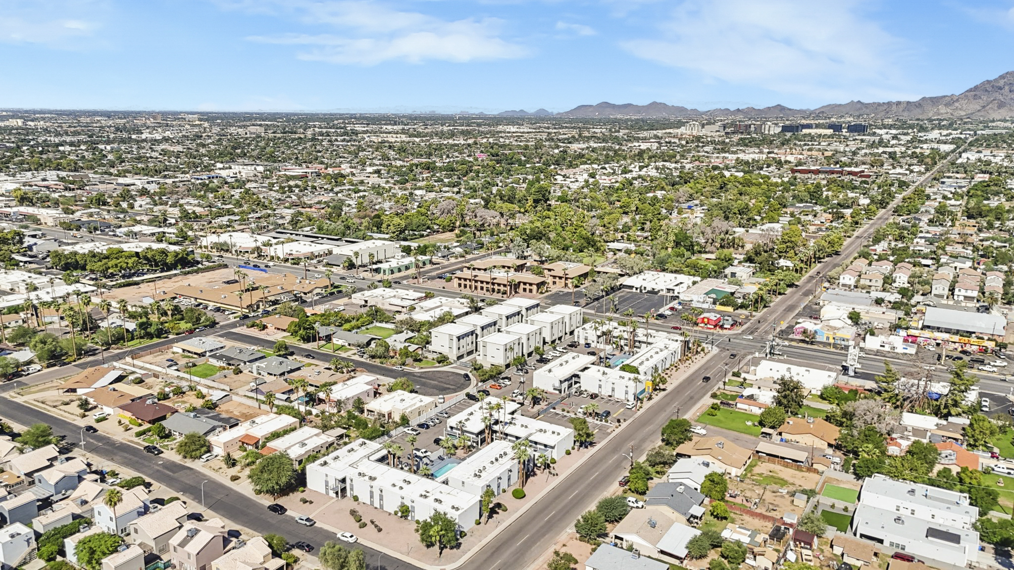 Aerial view of a suburban area featuring a mix of residential homes, commercial buildings, and green spaces. The landscape includes streets, parking lots, and various types of architecture, with distant mountains visible under a clear blue sky.