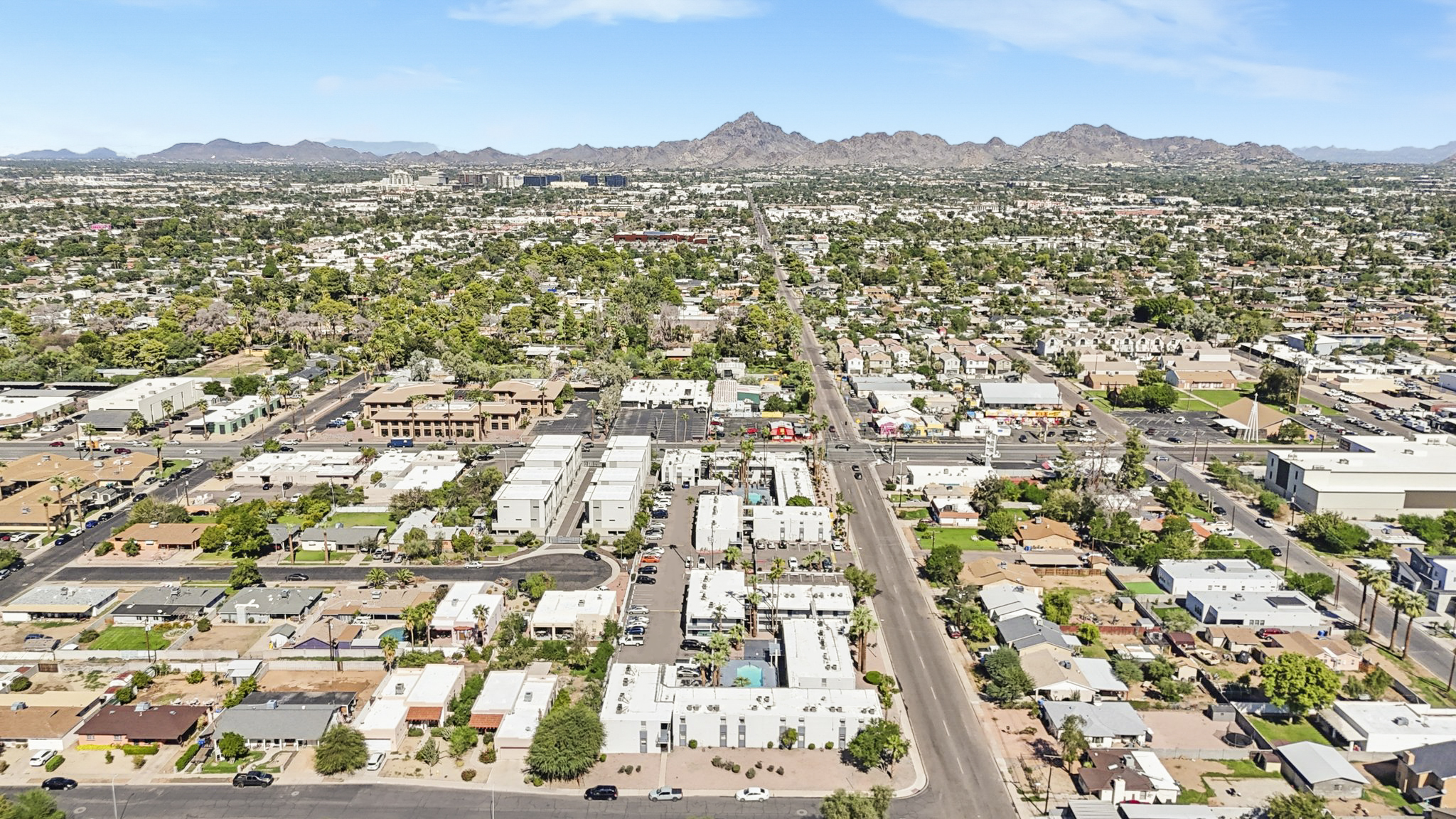 Aerial view of a suburban area featuring residential homes, commercial buildings, and roads, with mountains in the background and clear blue skies. The landscape is densely populated with greenery and various structures, illustrating a mix of urban and natural environments.