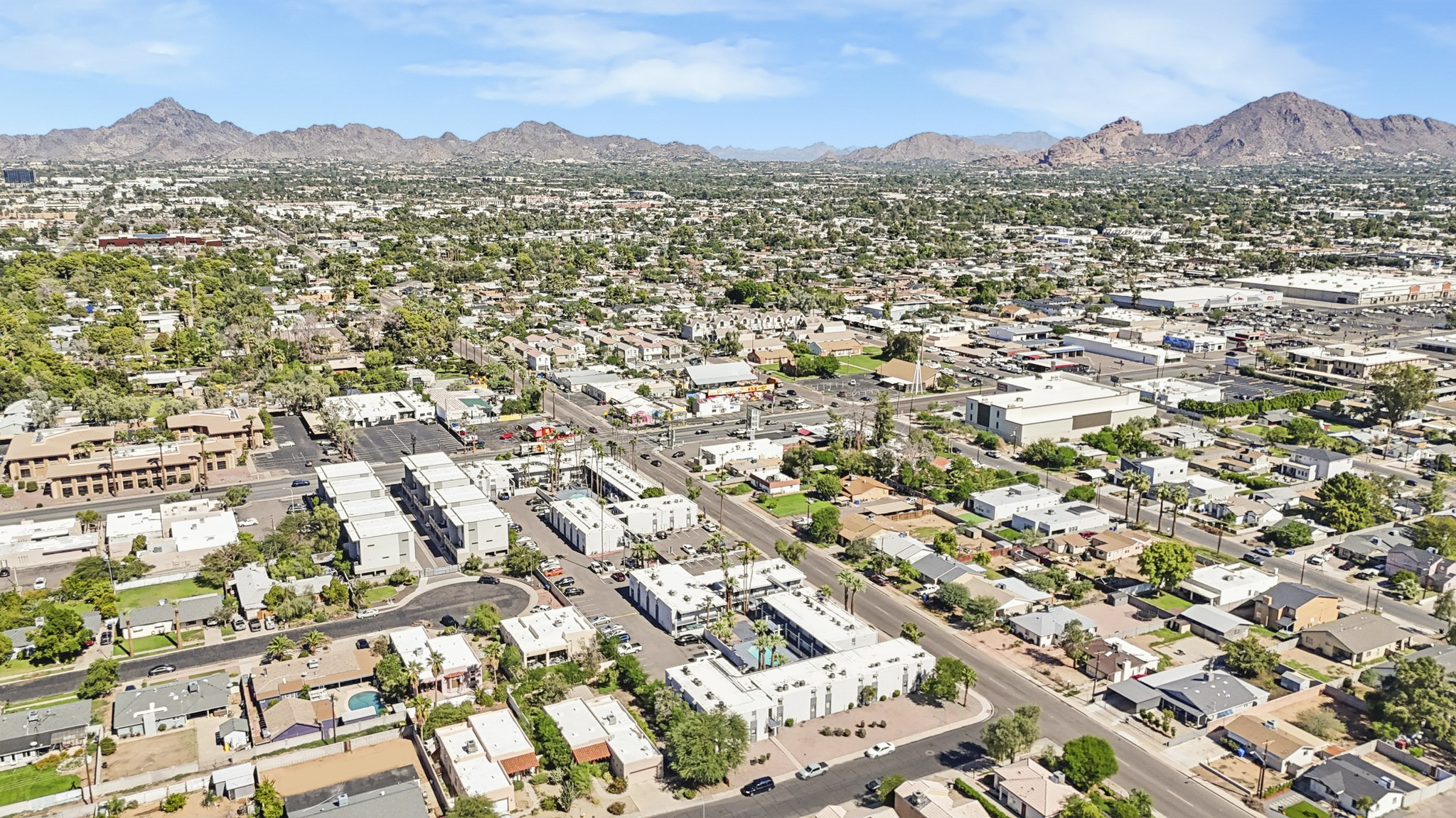 Aerial view of a suburban area with a mix of residential homes and commercial buildings. The landscape includes streets, trees, and a backdrop of mountains under a clear blue sky. The scene showcases the development and layout of the neighborhood, with various structures aligned along the roads.