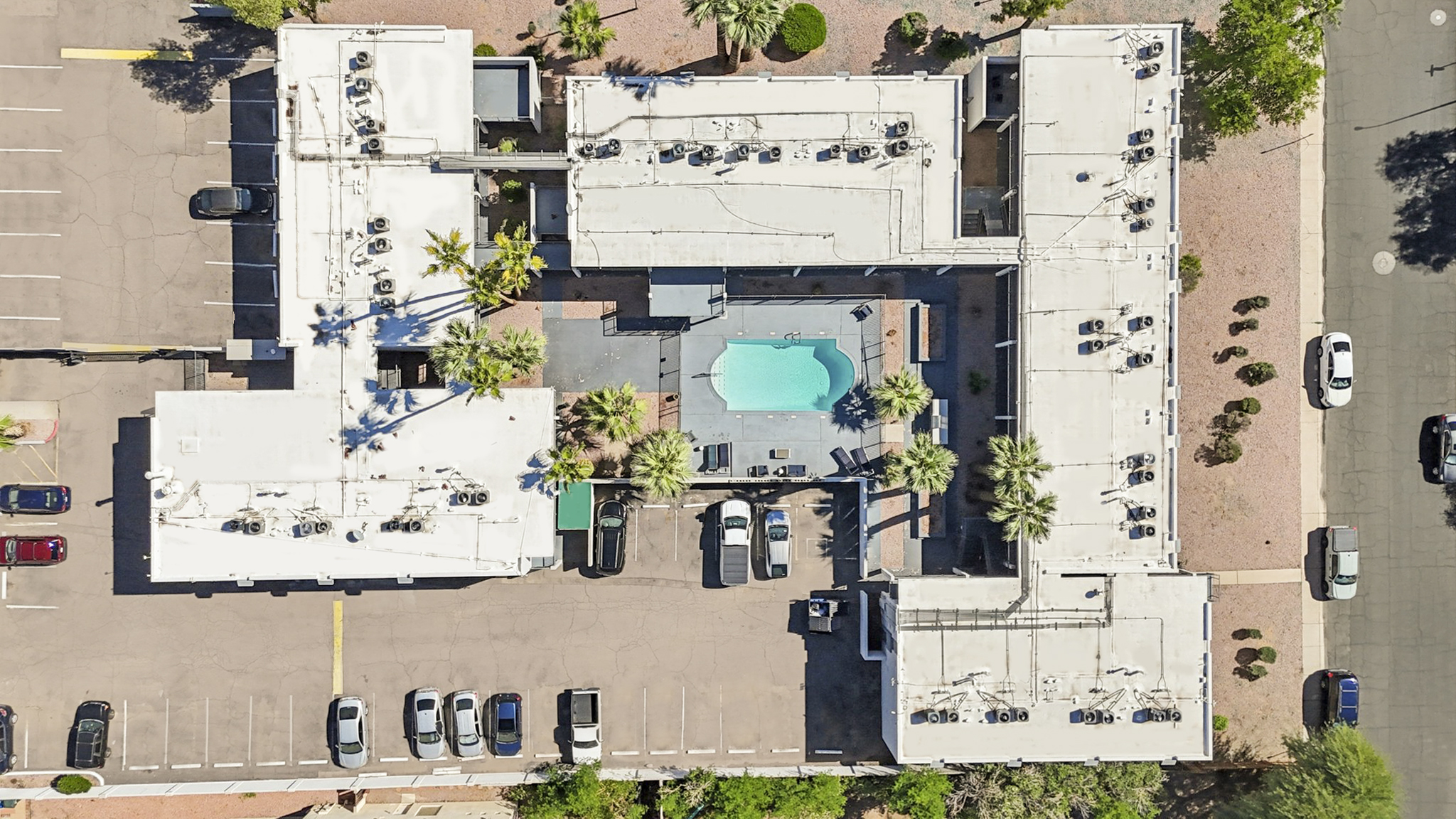 An overhead view of a hotel complex featuring several buildings arranged around a central pool area. The pool is surrounded by palm trees, and there are parking spaces filled with vehicles nearby. The buildings have flat roofs with visible air conditioning units. The area is well-maintained with landscaped surroundings.