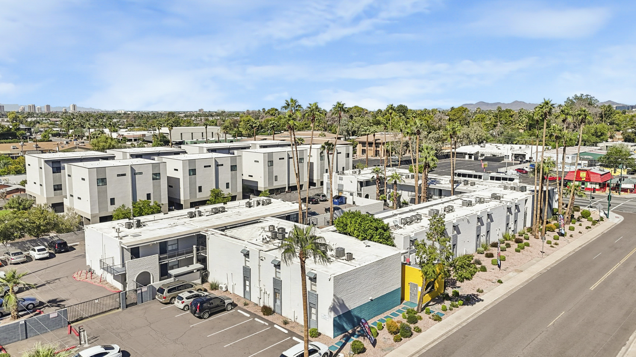 Aerial view of a commercial area featuring a low-rise building with multiple air conditioning units on the roof, surrounded by palm trees. The foreground shows a parking lot with several parked cars, while additional buildings and greenery are visible in the background against a blue sky.