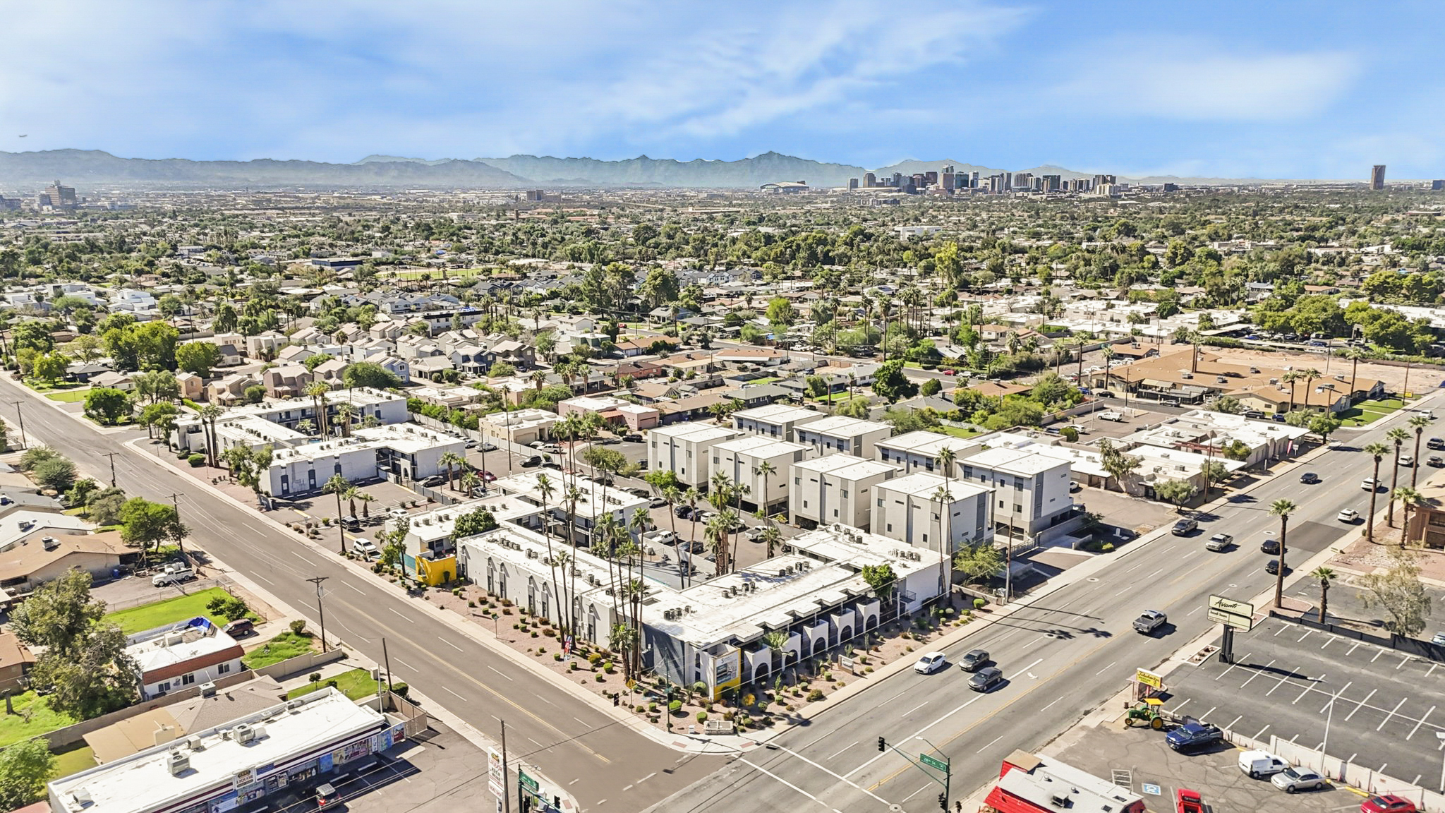 Aerial view of a suburban area featuring low-rise buildings, palm trees, and various commercial and residential structures. In the background, a skyline with tall buildings is visible against a clear blue sky, with mountains in the distance. The scene captures a bustling urban environment.