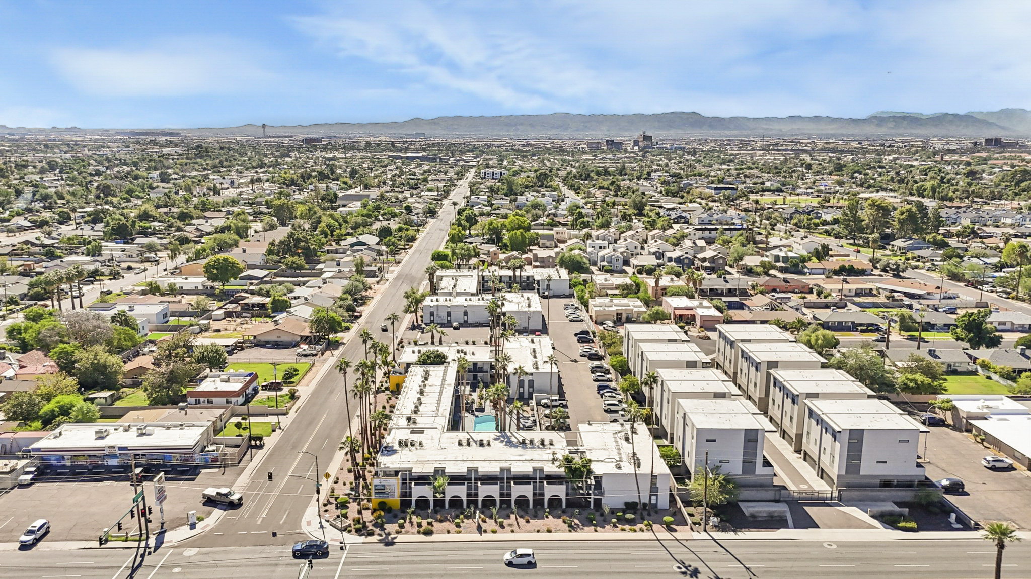 Aerial view of a suburban area featuring a mix of residential and commercial buildings, tree-lined streets, and a clear blue sky. The image shows multiple housing units and a swimming pool area, with mountains visible in the distance.