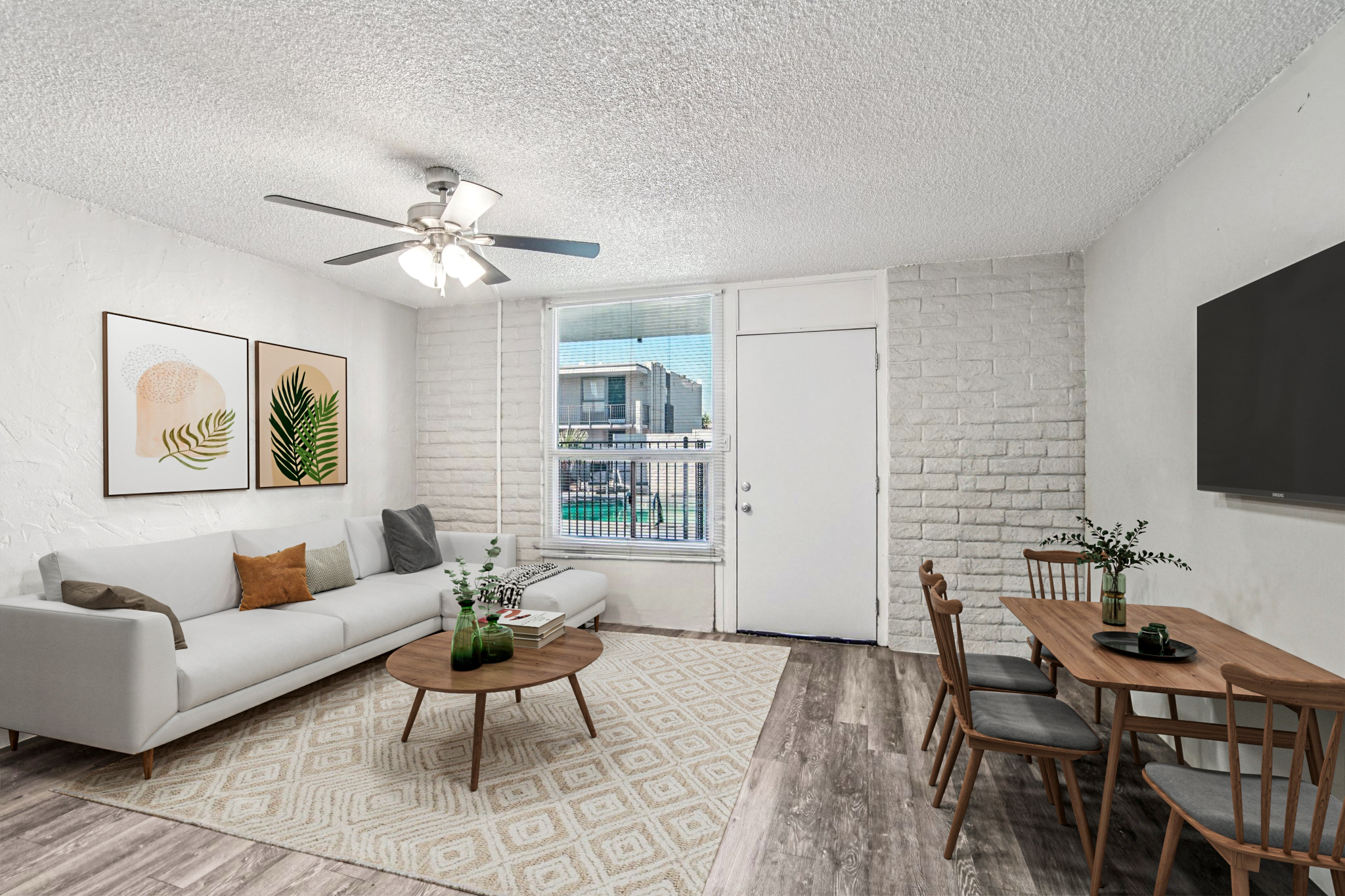 A bright and inviting living room featuring a light gray sofa with decorative pillows, a round coffee table, and a dining table with wooden chairs. The walls have modern artwork of plants, and large windows allow natural light in, showcasing a view of the outdoors.