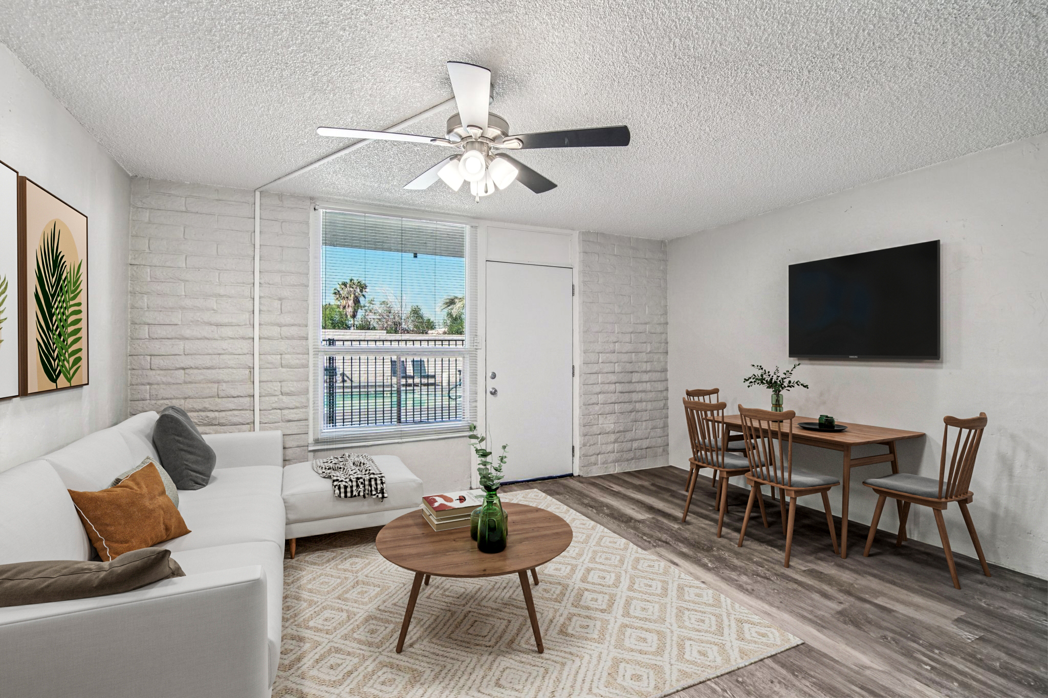 A modern living room with a white sofa, wooden dining table, and chairs. A ceiling fan is visible, along with a wall-mounted TV. The room features a large window with a view of palm trees outside, light wood flooring, and decorative plants in a vase on the table. The overall decor is bright and inviting.