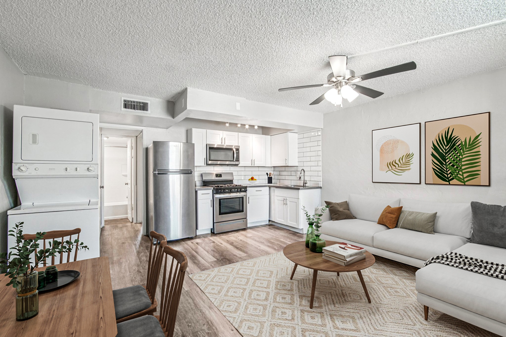 A modern living space featuring an open kitchen and living room layout. The kitchen has stainless steel appliances, including a refrigerator and an oven. There's a comfortable white sofa, a round coffee table with books and a plant, and a dining area with wooden chairs. Two framed botanical prints adorn the wall.