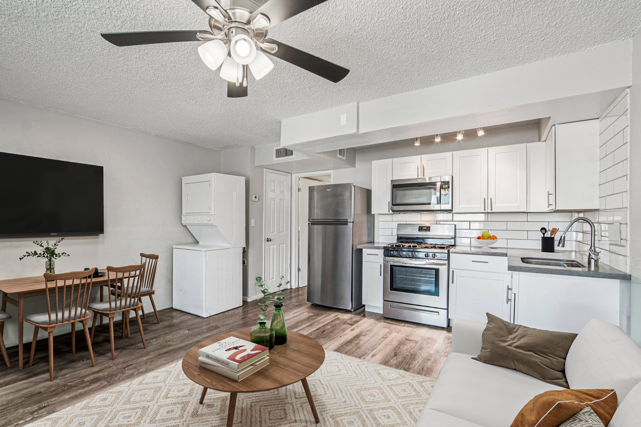 A modern kitchen and living area featuring a stainless steel refrigerator, stove, and microwave. A dining table with wooden chairs is nearby, and a cozy white sofa with decorative pillows is in the foreground. The space has a light color scheme and a ceiling fan, creating a bright and inviting atmosphere.